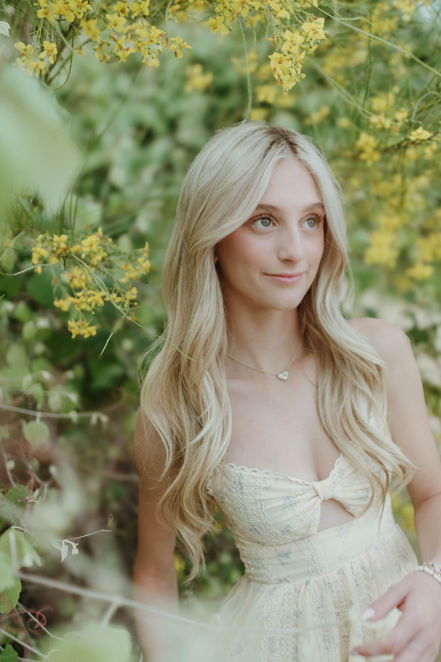 A blonde woman in a cream dress smiles softly surrounded by yellow flowering greenery.