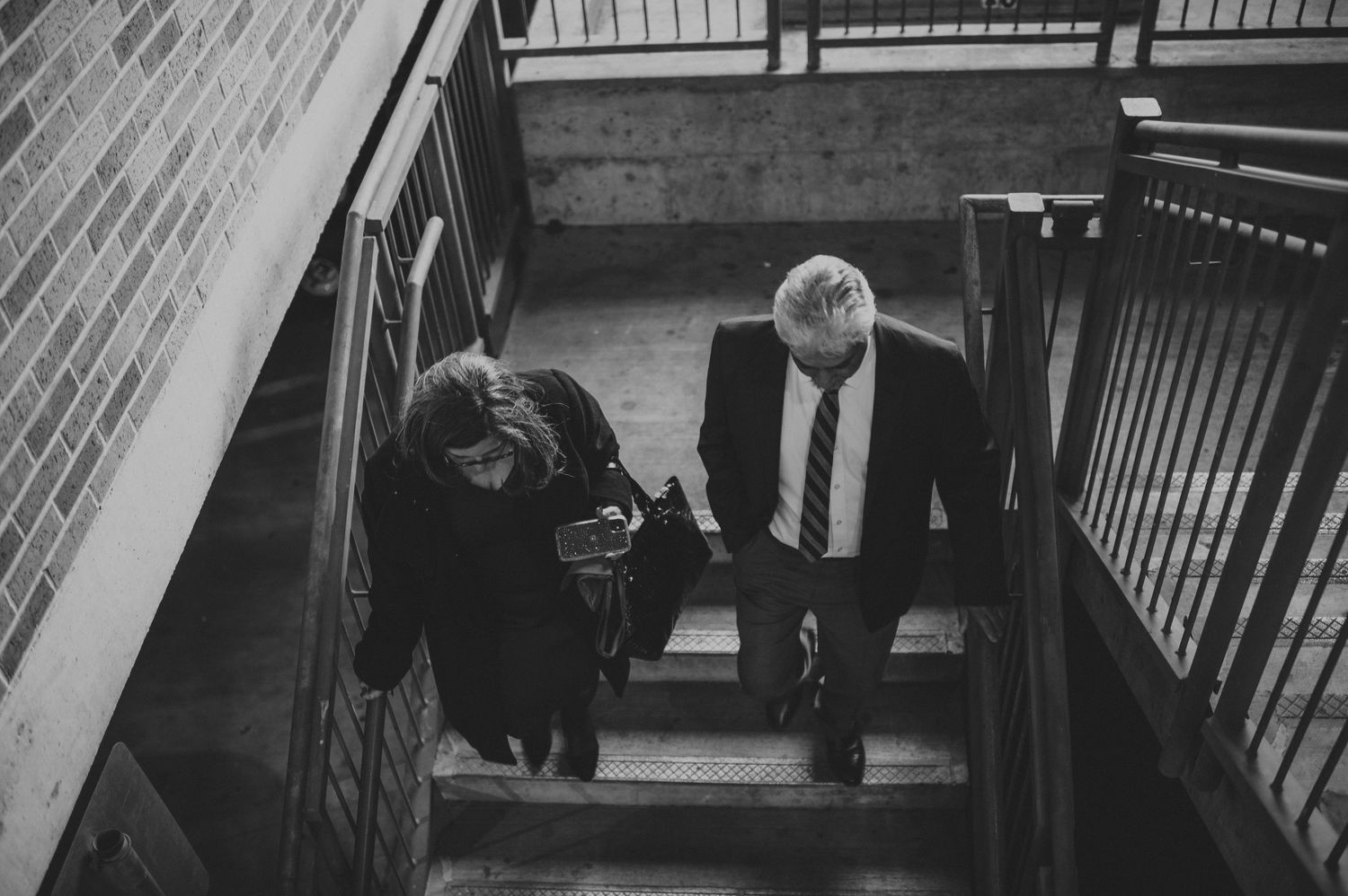 Aerial black and white view of two people ascending interior brick building staircase.