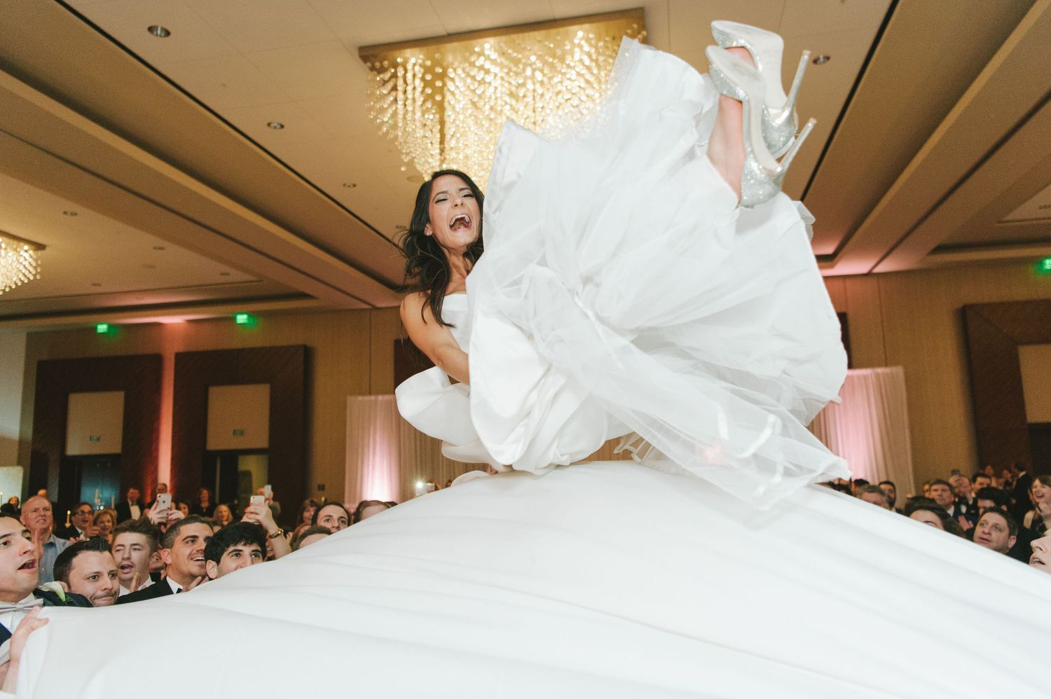 Joyful bride being lifted in the air in her white wedding gown during a lively reception celebration.
