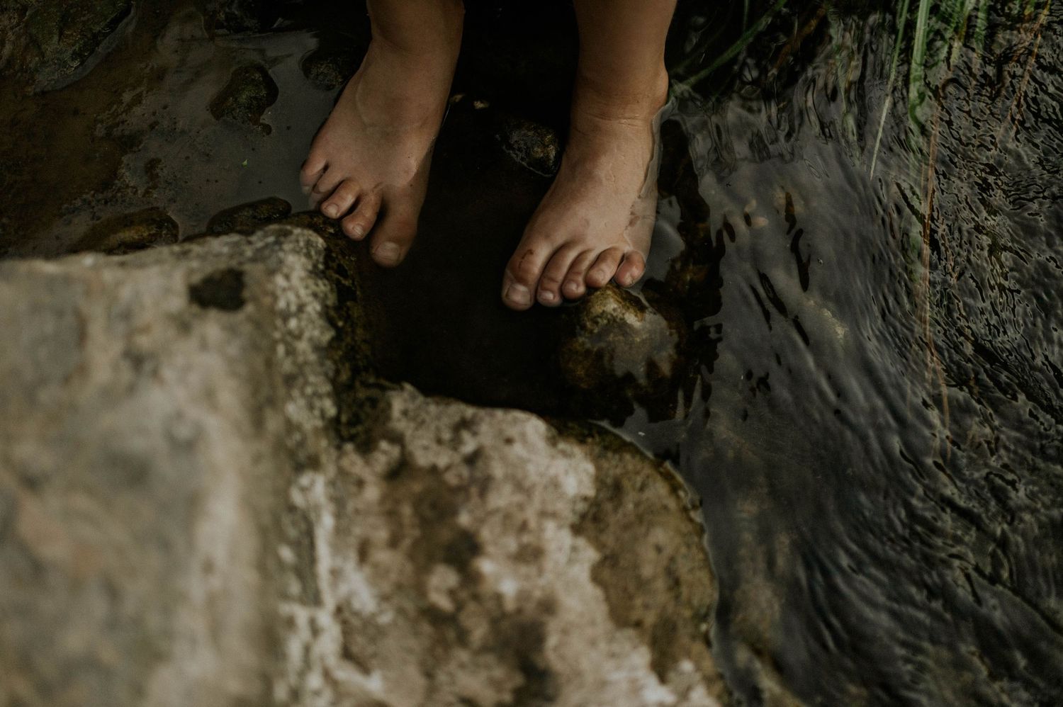 Bare feet of a child standing on a mossy rock beside a flowing stream in nature.