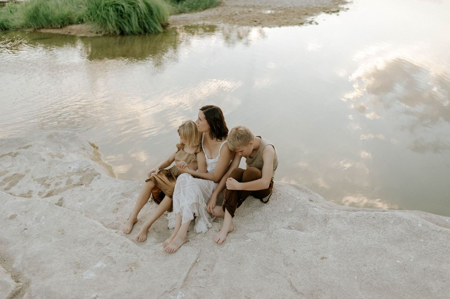 A mother and two children share a close embrace while sitting on rocks beside a calm river.