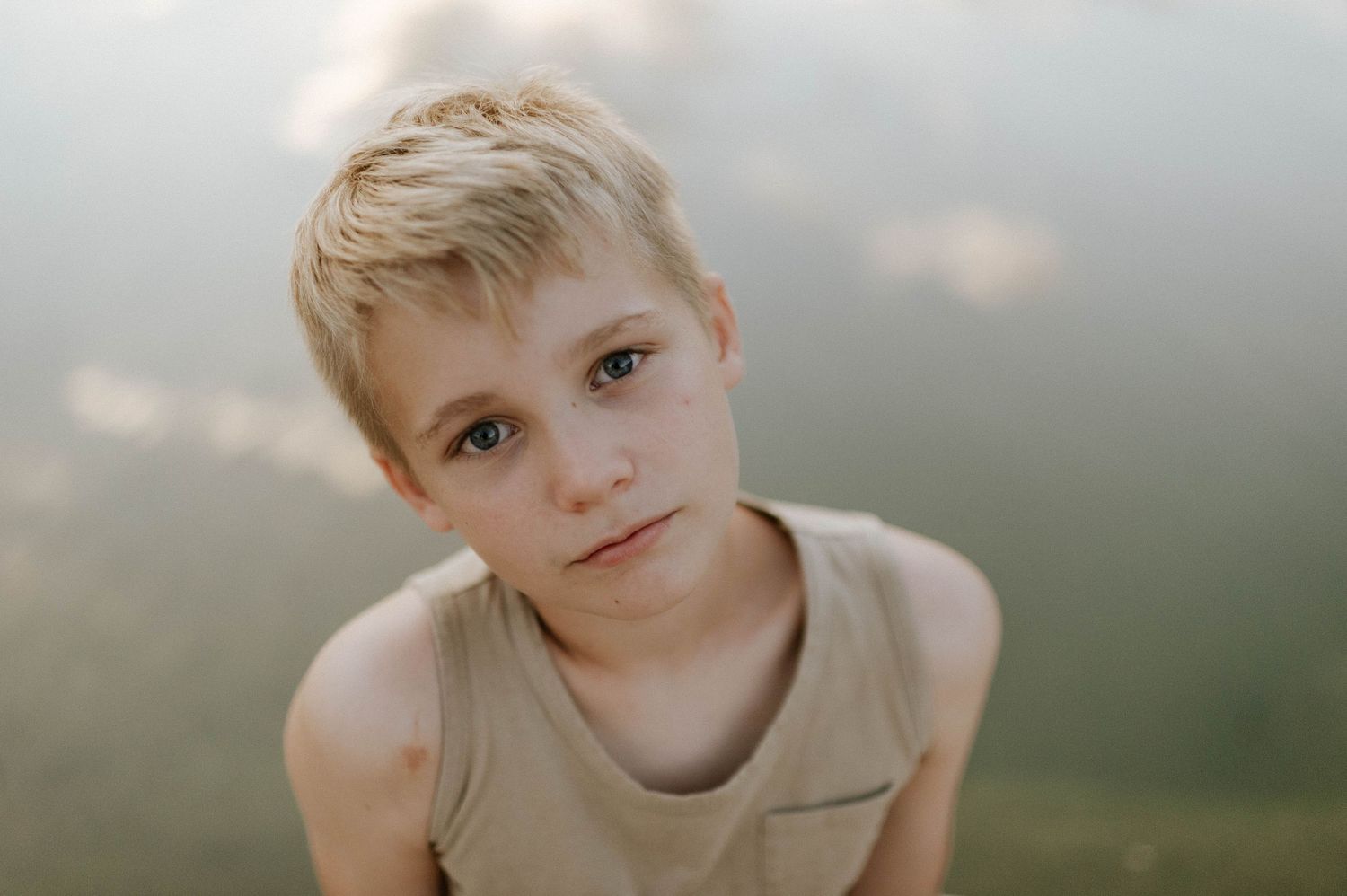 Serious young blonde boy in a tan tank top sitting near a misty lake.