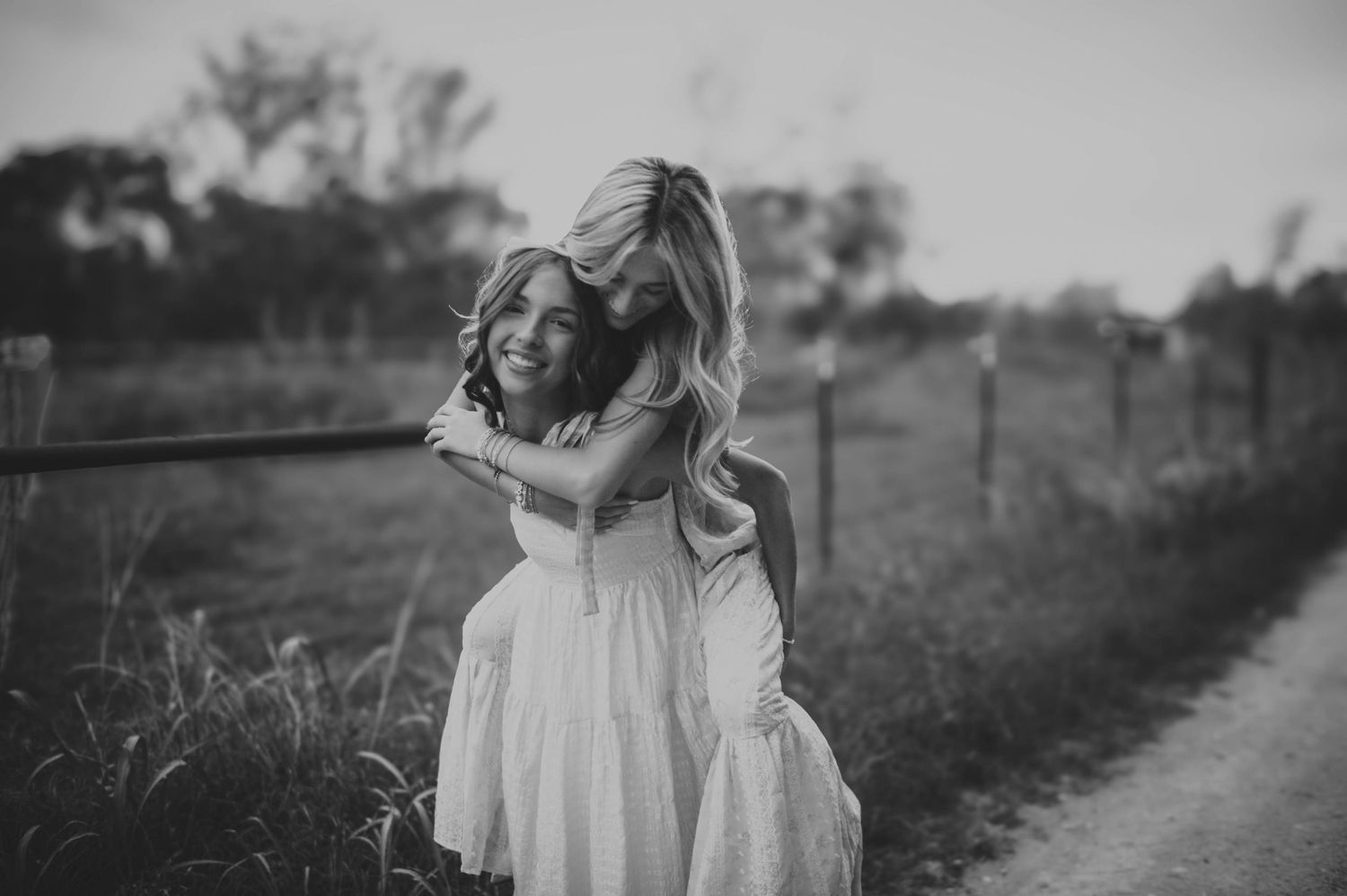 Two girls in white dresses share a joyful piggyback ride along a rural path.