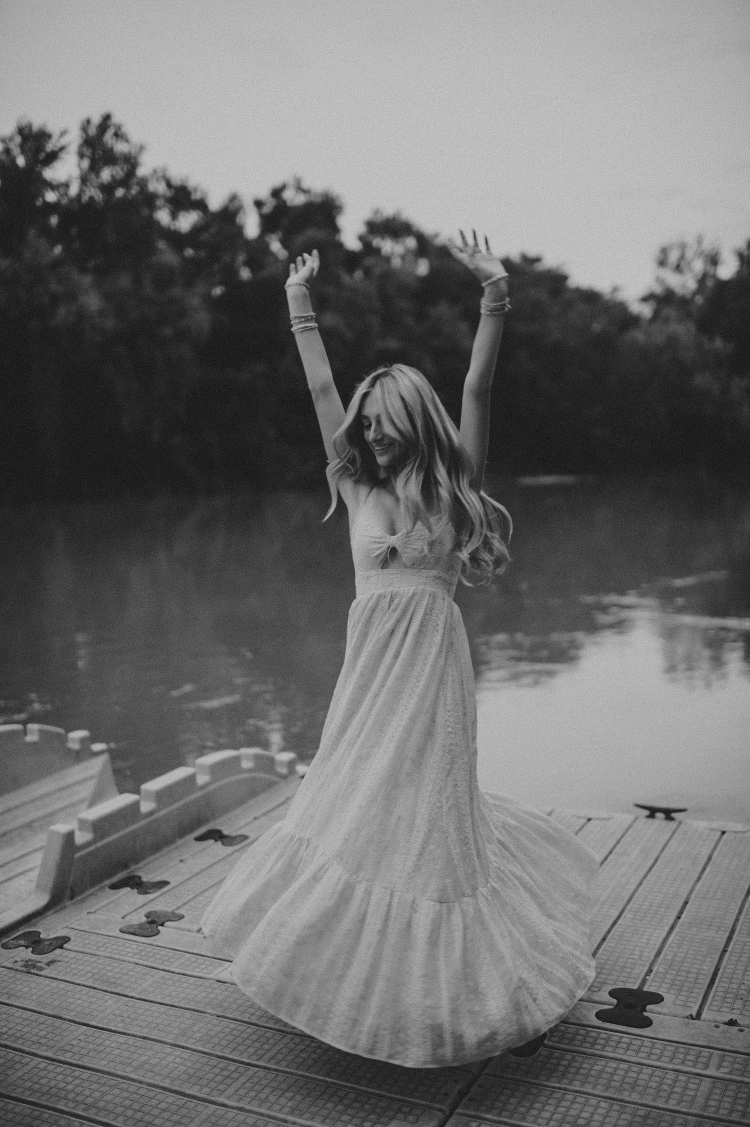 Woman in a flowing white dress dancing joyfully on a lakeside dock with arms raised overhead.