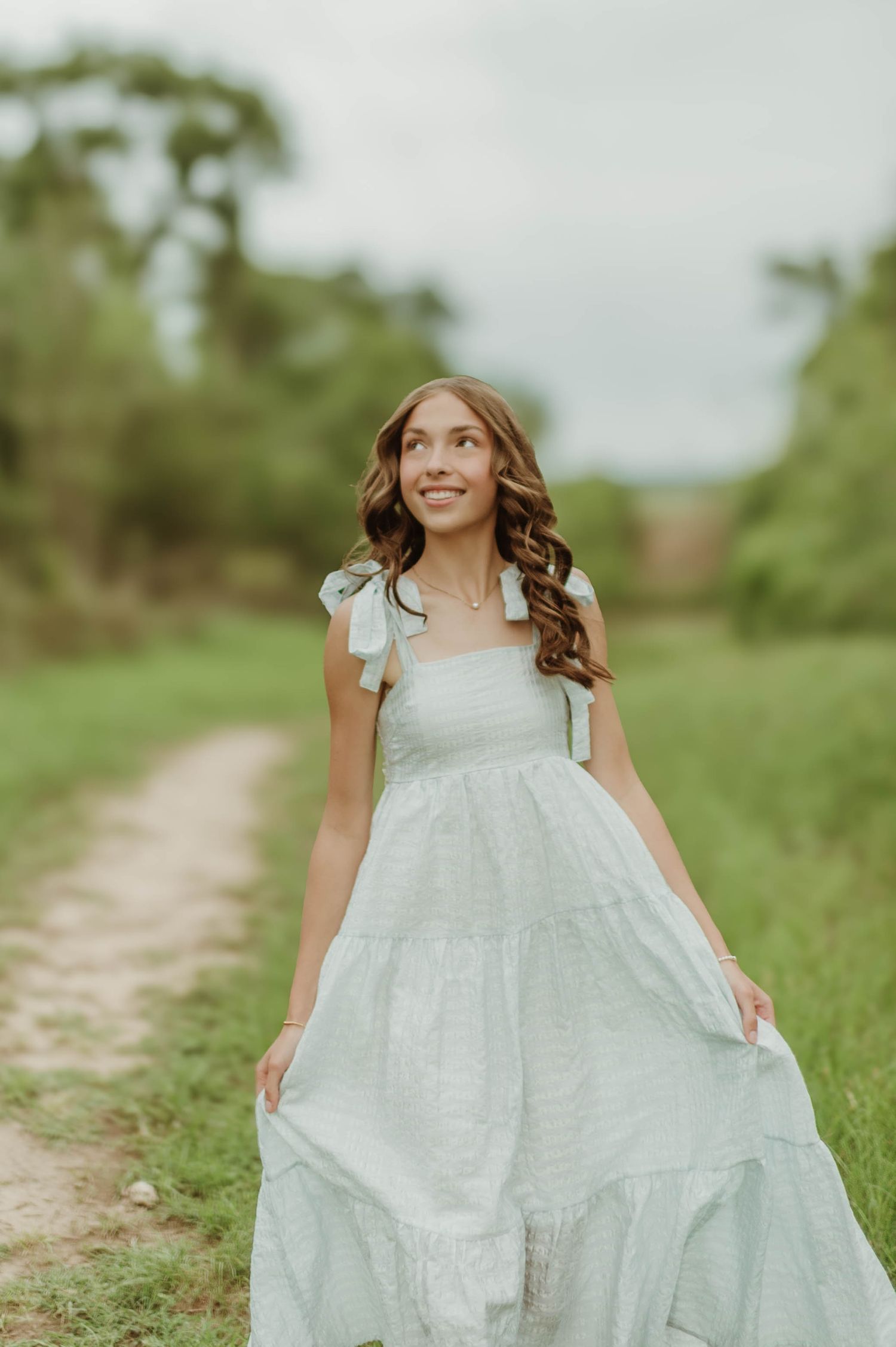 Young girl in light blue smocked maxi dress walking along a dirt path in a green outdoor landscape.