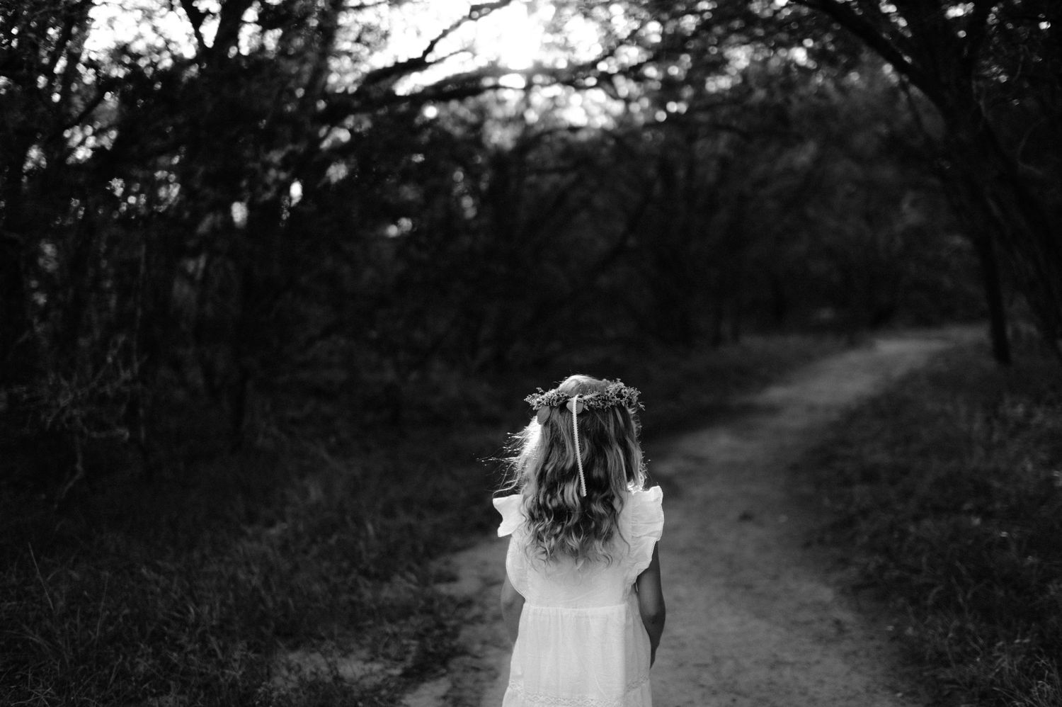 Young girl in white dress with floral crown walks alone down a serene forest path in black and white.