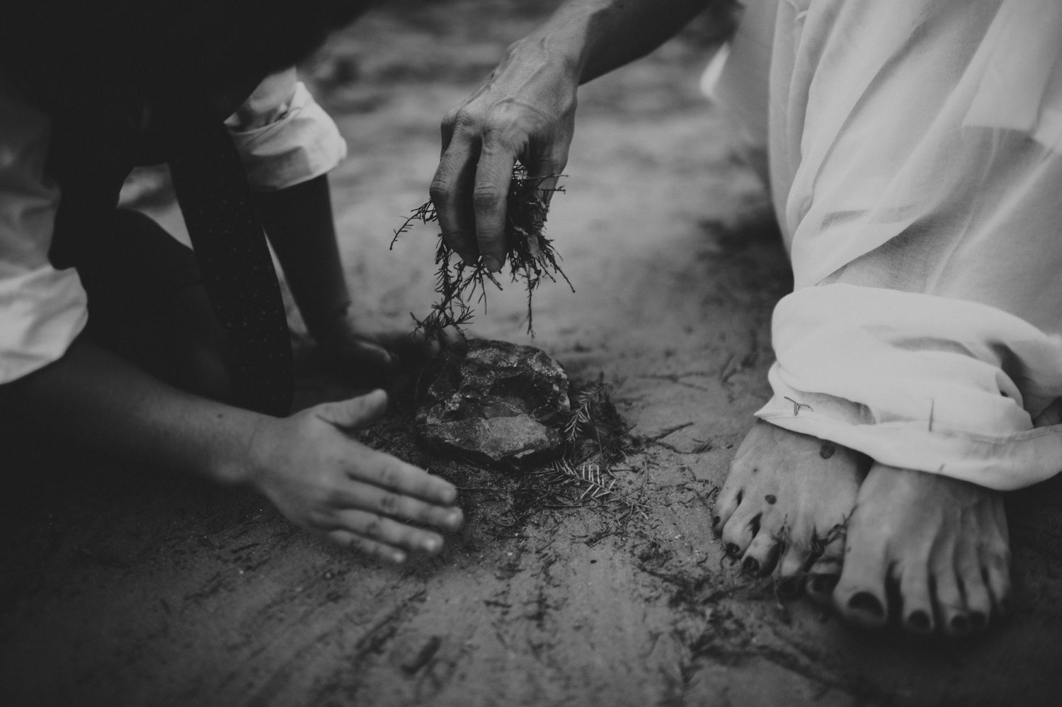 Black and white photo of hands placing a crown of thorns on sandy ground near bare feet.