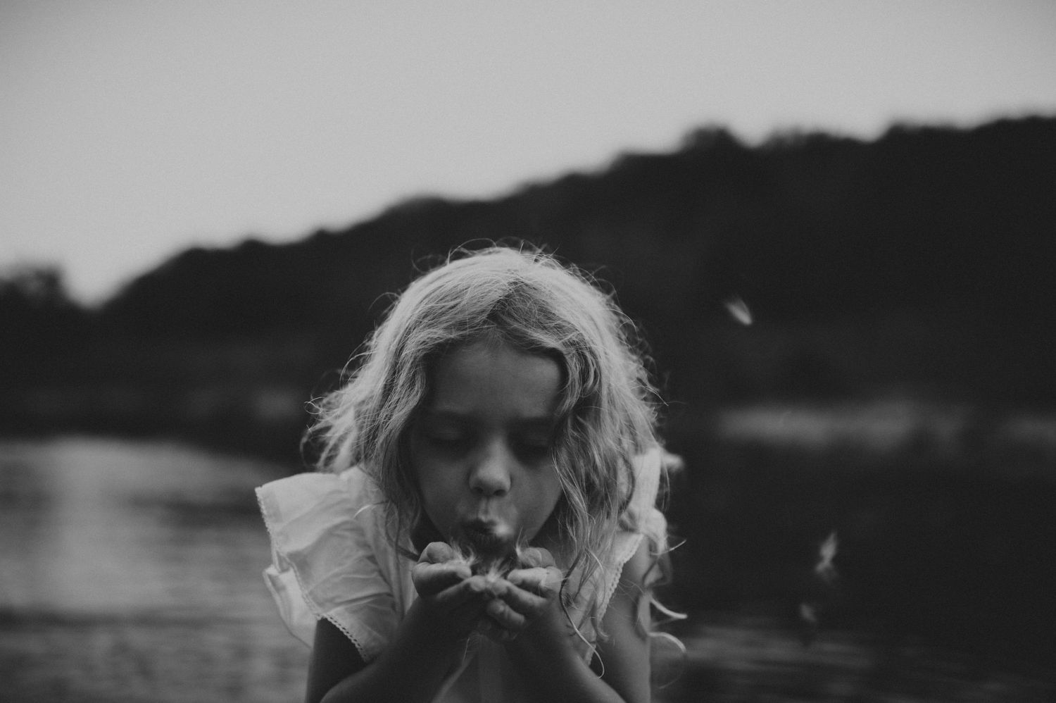 Black and white photo of a young blonde girl blowing dandelion seeds near a lake.