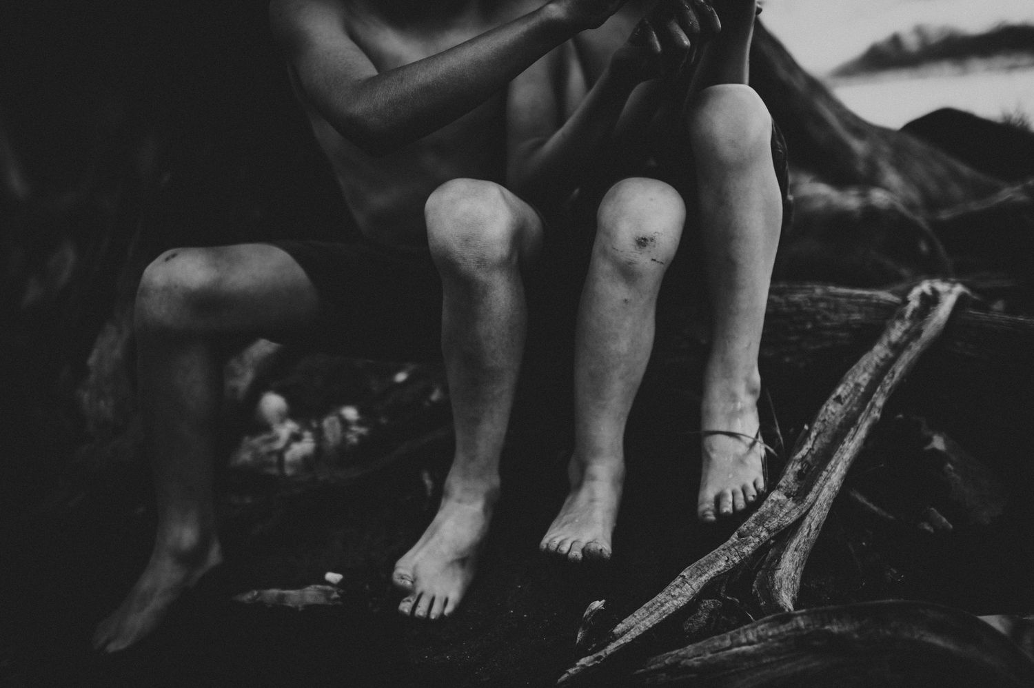 Black and white photo of two people sitting barefoot on driftwood, legs visible in an outdoor natural setting.