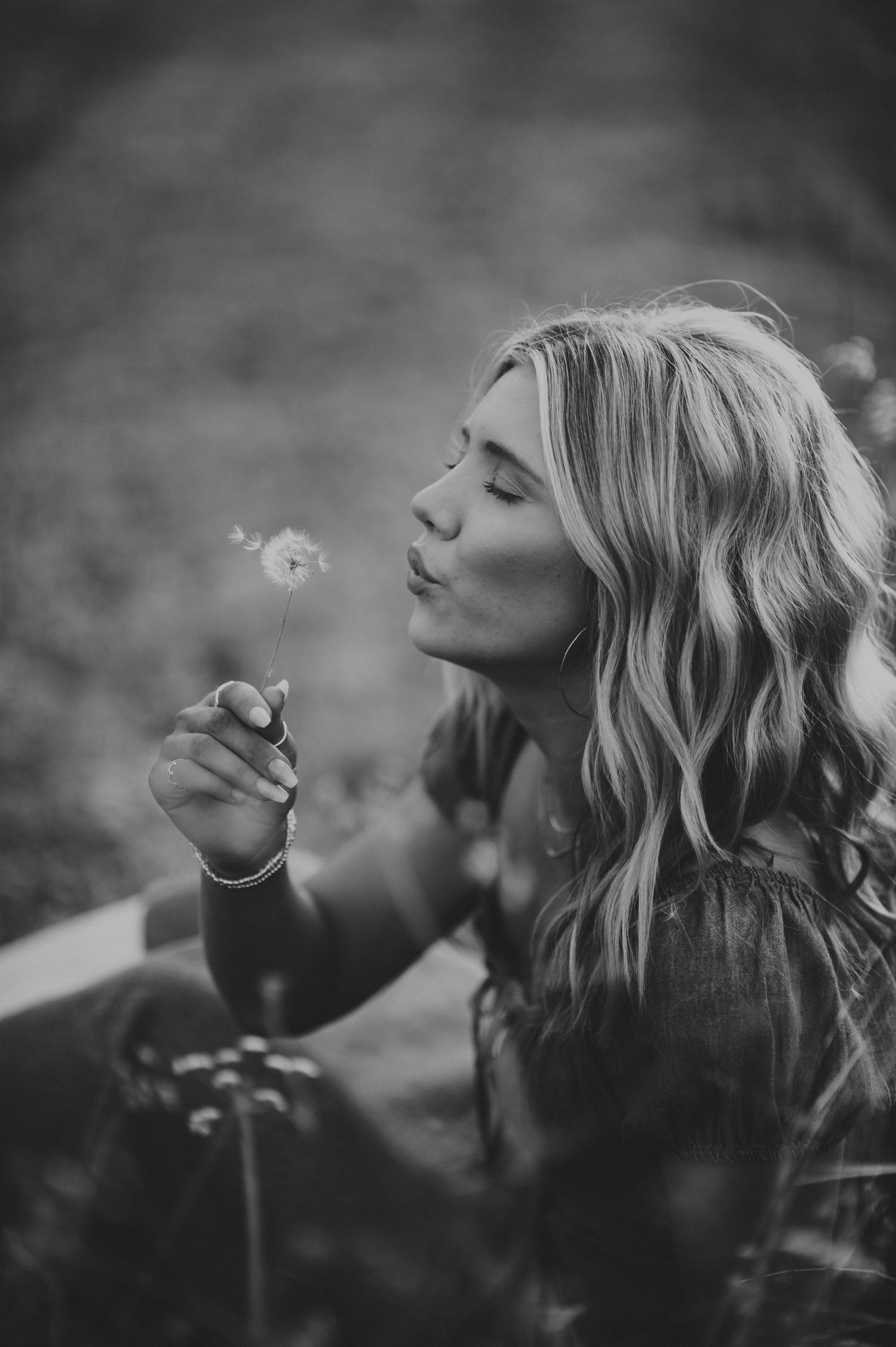 Black and white photo of a young woman with wavy blonde hair blowing a dandelion outdoors.