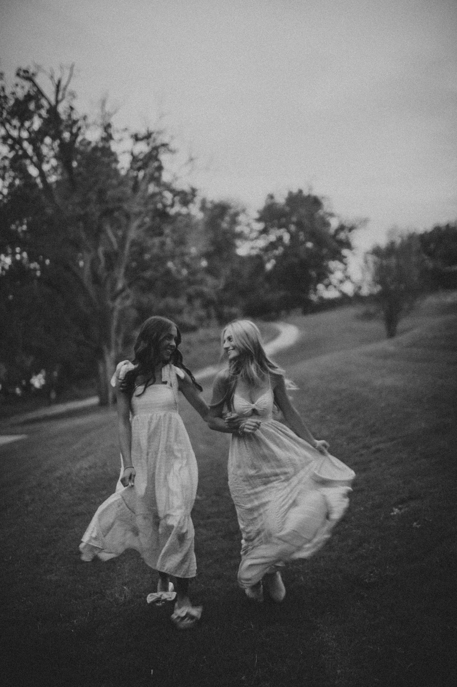 Two young women in white dresses twirling and holding hands on a grassy hill at dusk.