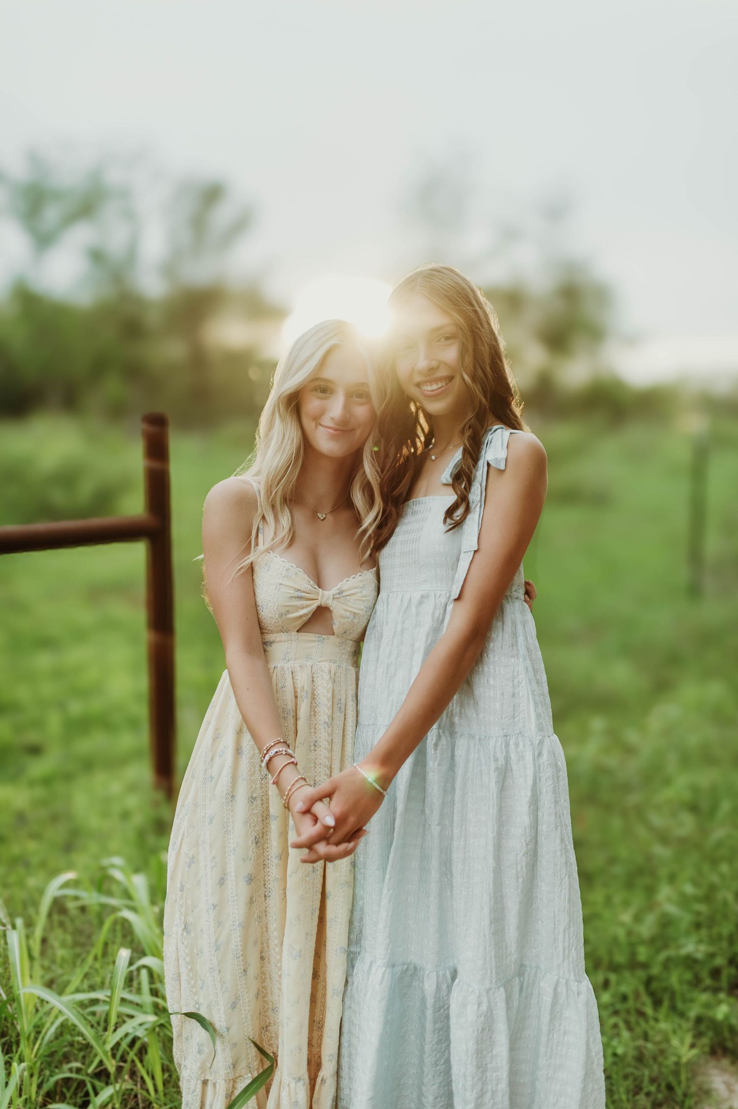 Two teenage girls in boho dresses holding hands with golden sunset glowing behind them in a green field.