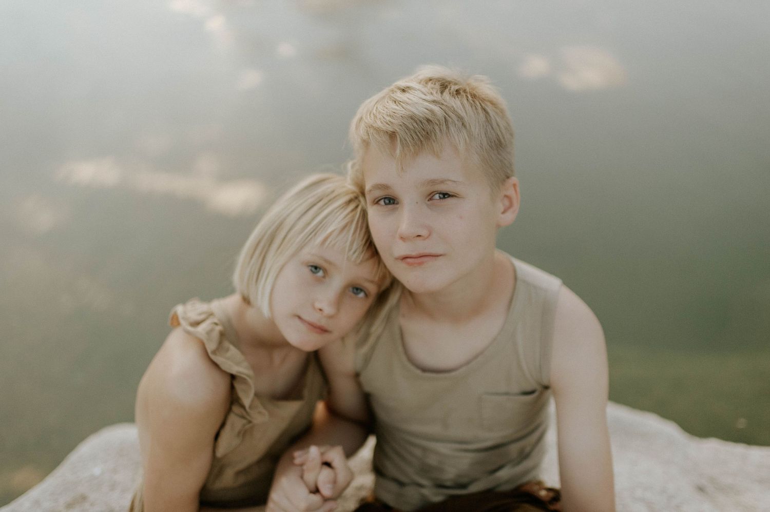Two blonde siblings hugging closely while sitting near a calm lake outdoors.