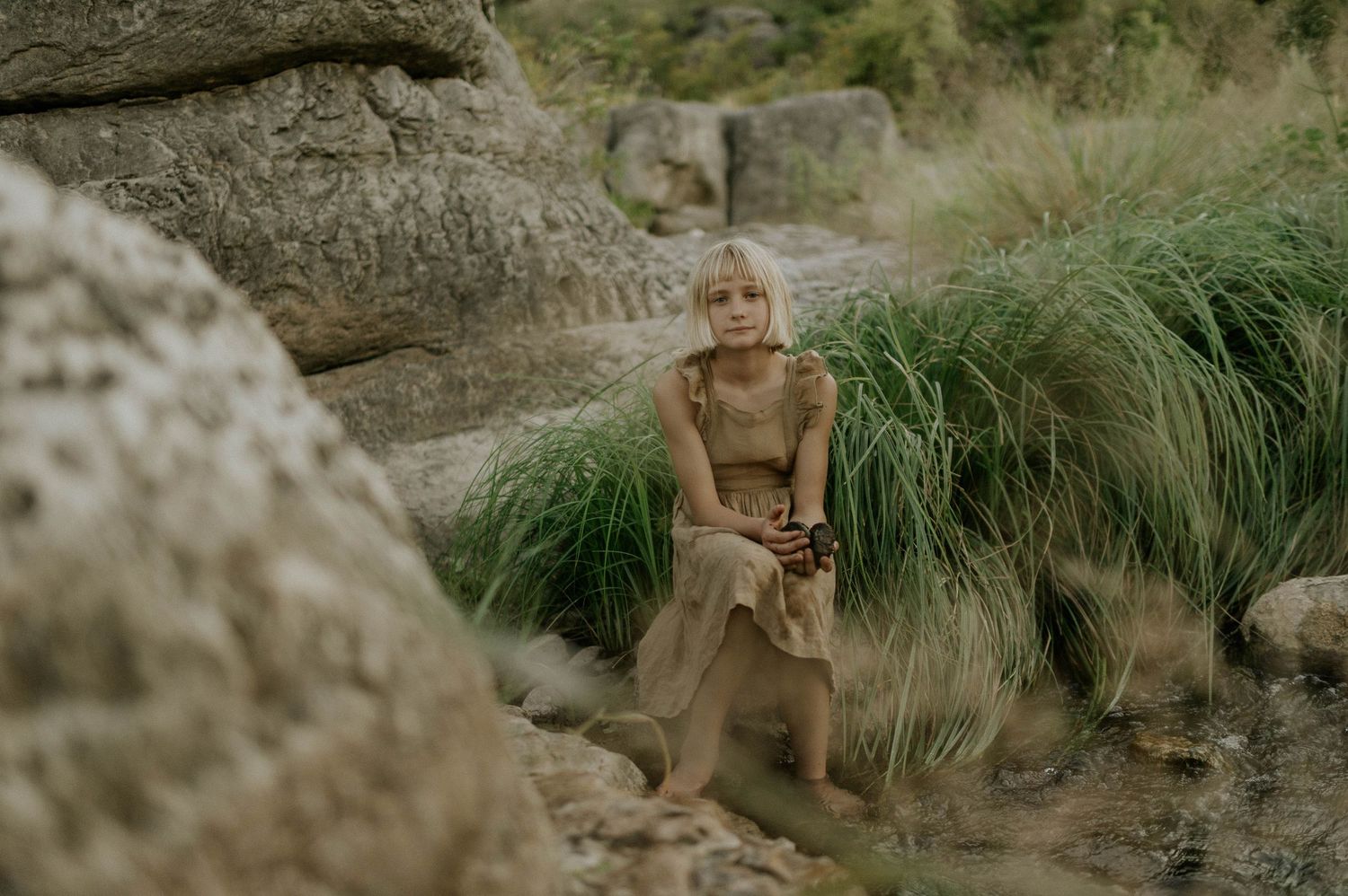Blonde girl in a tan dress sitting on rocks surrounded by tall green grass and boulders.