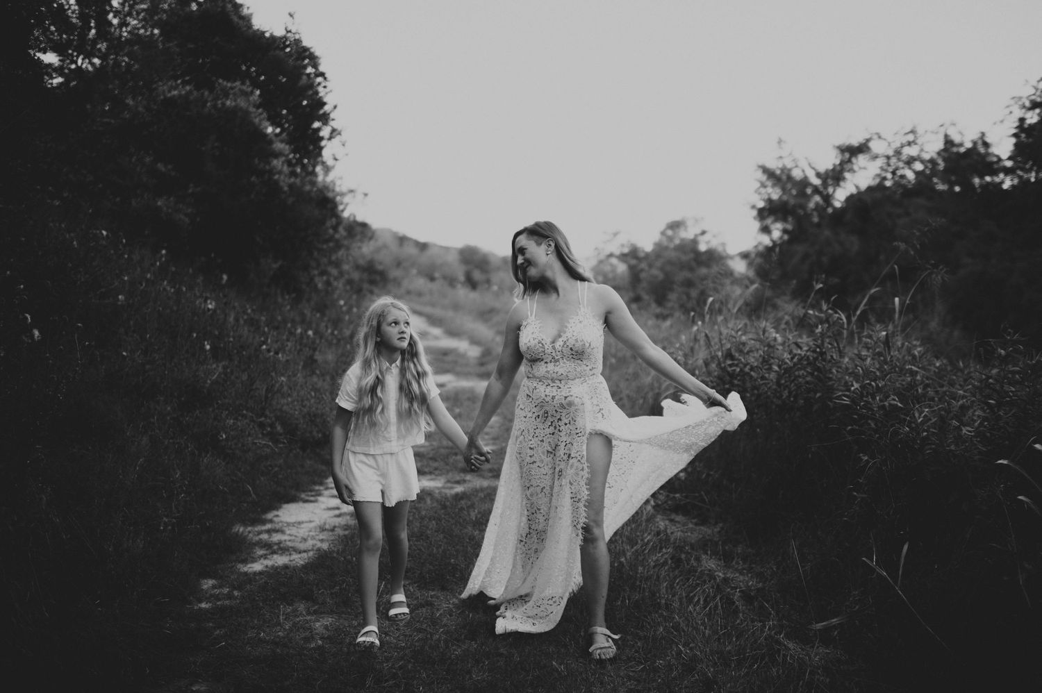 A woman in a flowing dress walks hand in hand with a young girl along a scenic nature path.