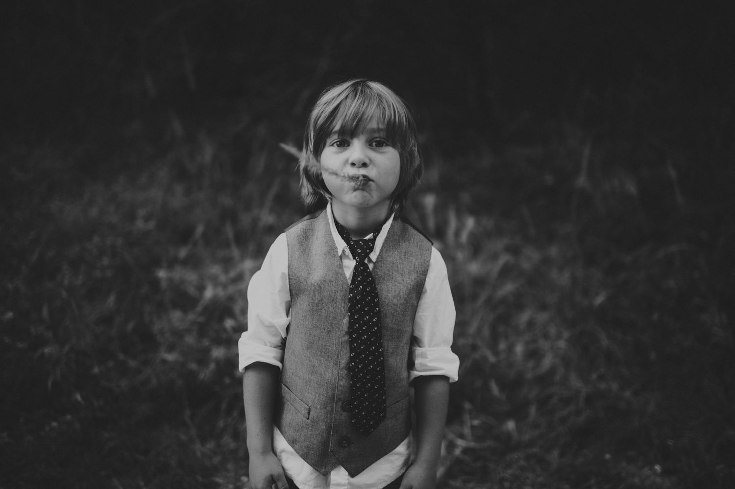 Black and white photo of a young boy in a vest and tie making a funny face outdoors.