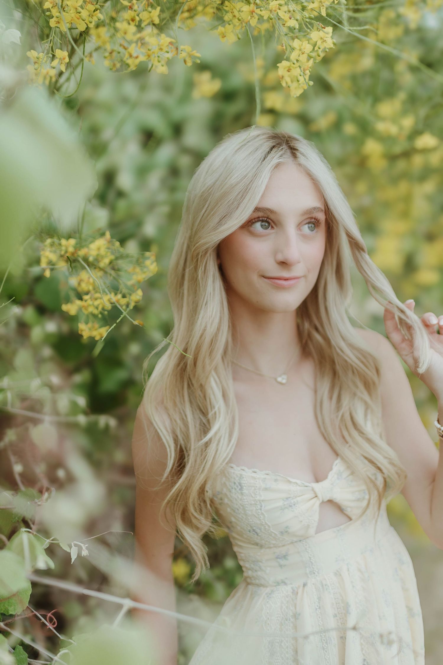Blonde teen girl in cream lace dress gazing softly among yellow wildflowers in a natural outdoor setting.