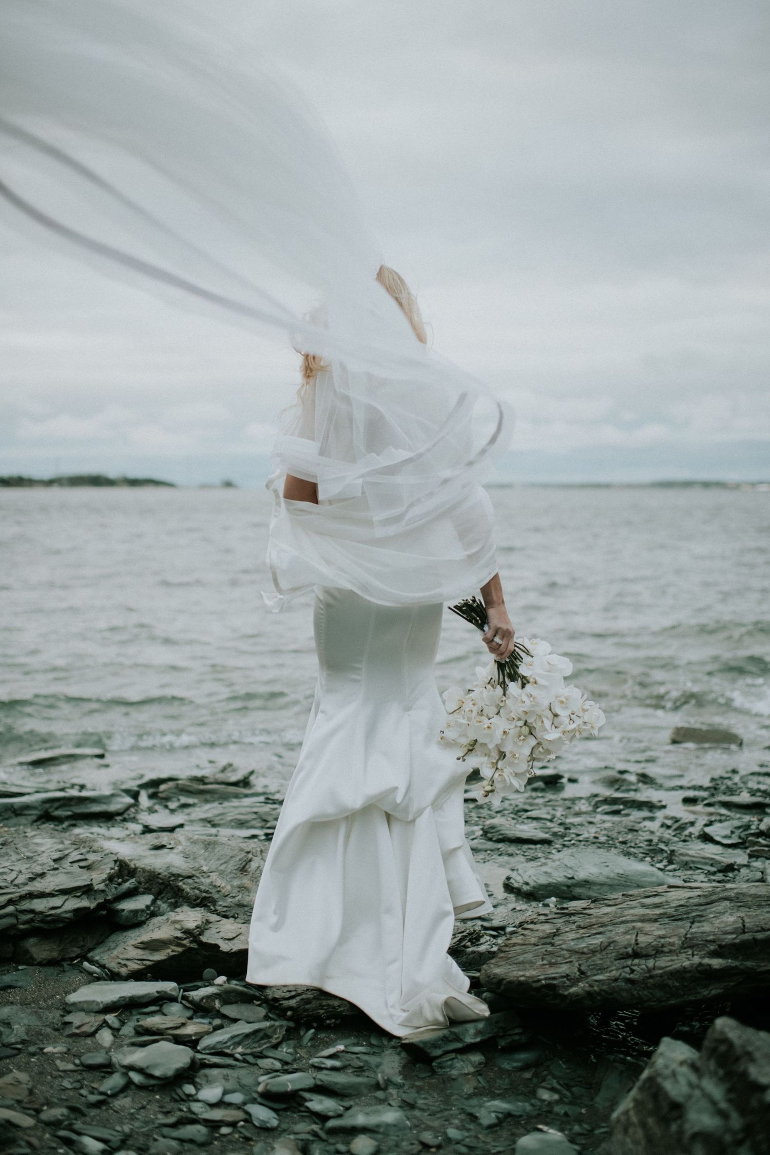 Brides veil billows dramatically in the wind as she stands on rocky shore holding white bouquet.