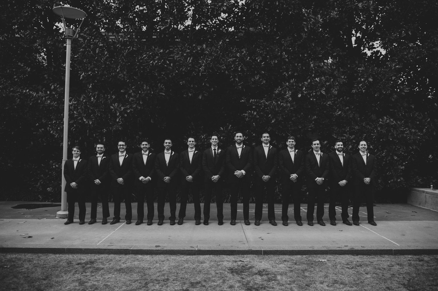 Black and white photo of groomsmen standing in a straight line wearing matching dark suits outdoors.