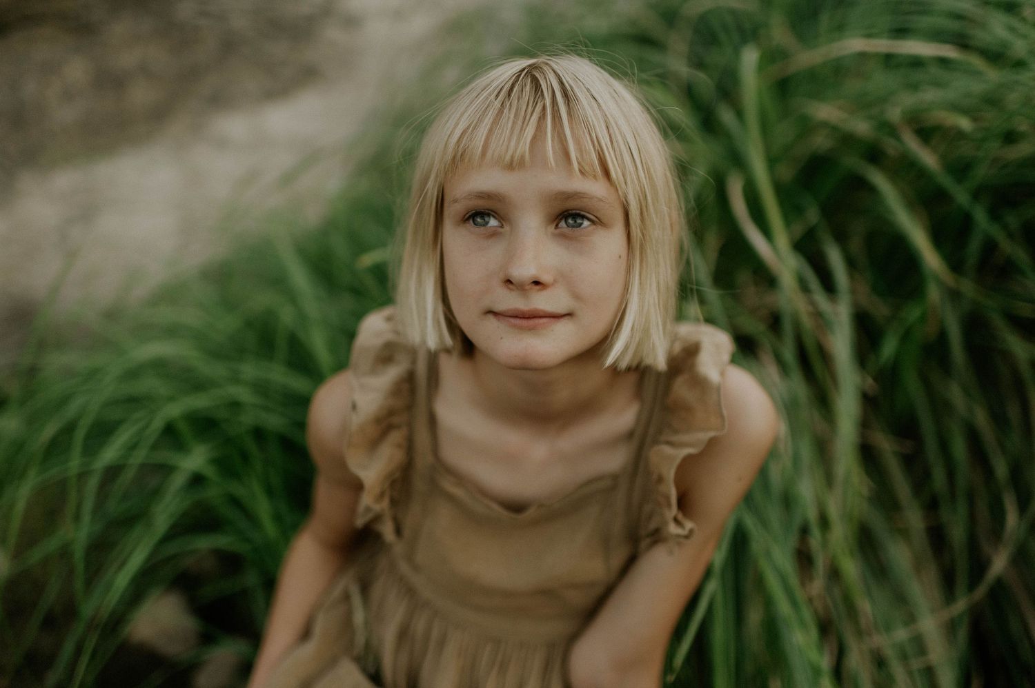 Young blonde girl with blue eyes wearing a tan dress sitting among tall green grass outdoors.