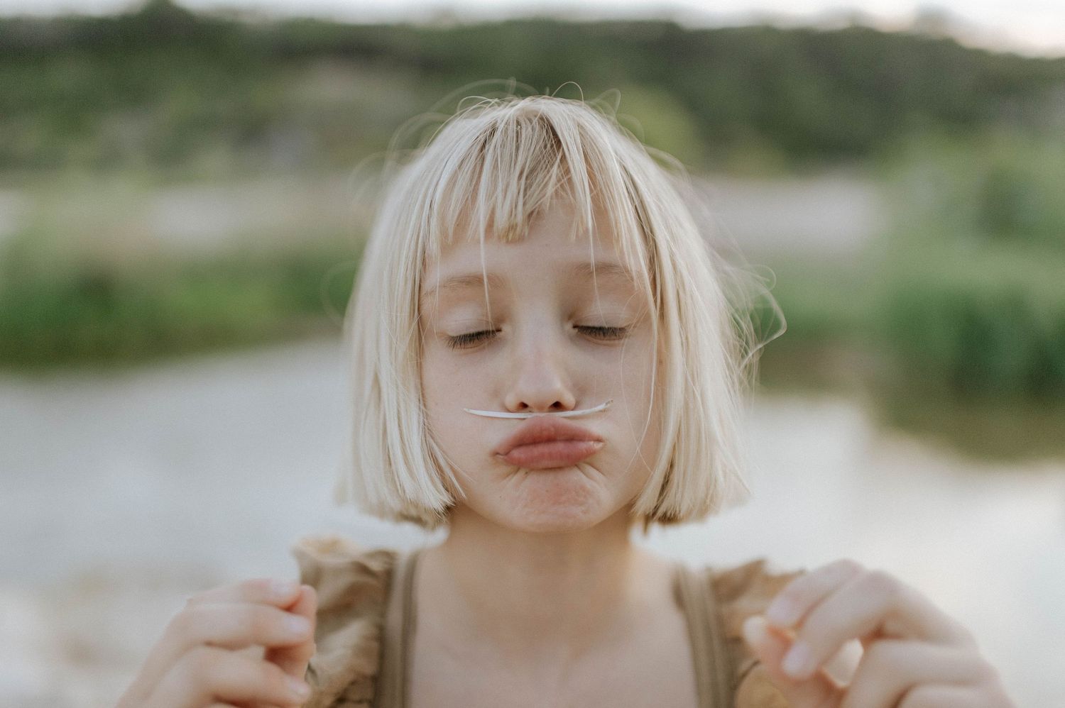 Blonde girl with bangs making a funny face with a plant stem near her lips outdoors.