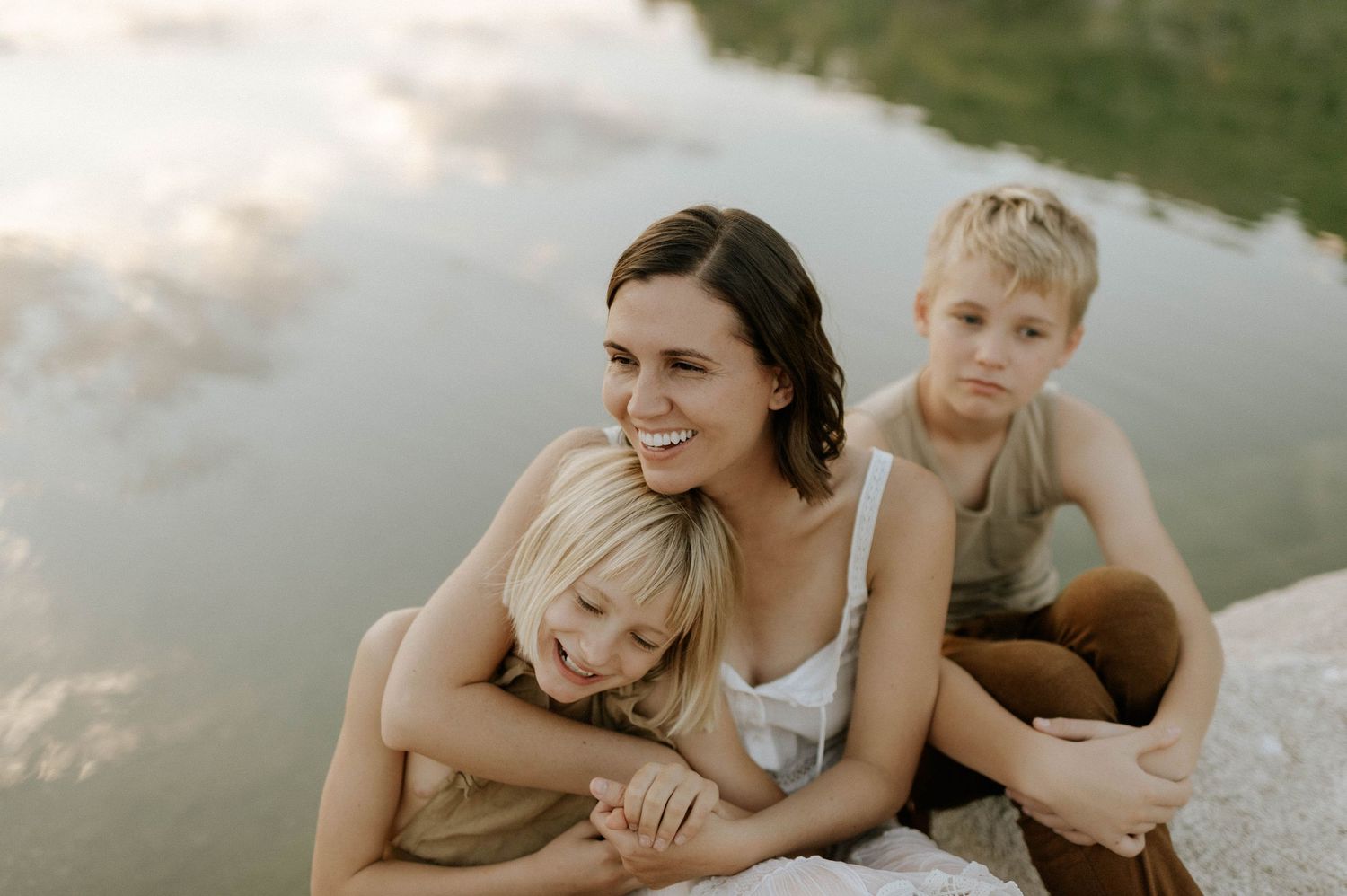 A smiling mother hugs her daughter while her son sits beside them on a rocky riverbank.