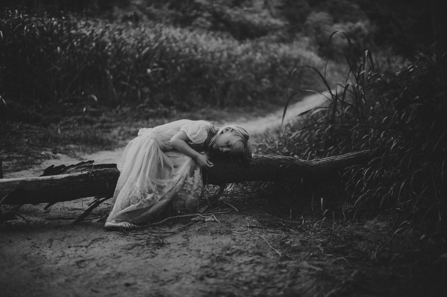 Black and white photo of a woman in a white dress lying on the ground in a dark, moody outdoor setting.