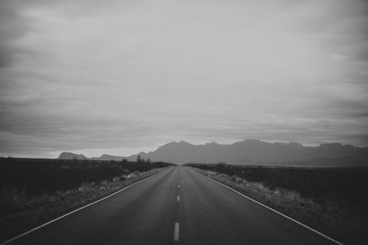 Desolate straight highway leading toward distant mountains under cloudy sky in black and white.