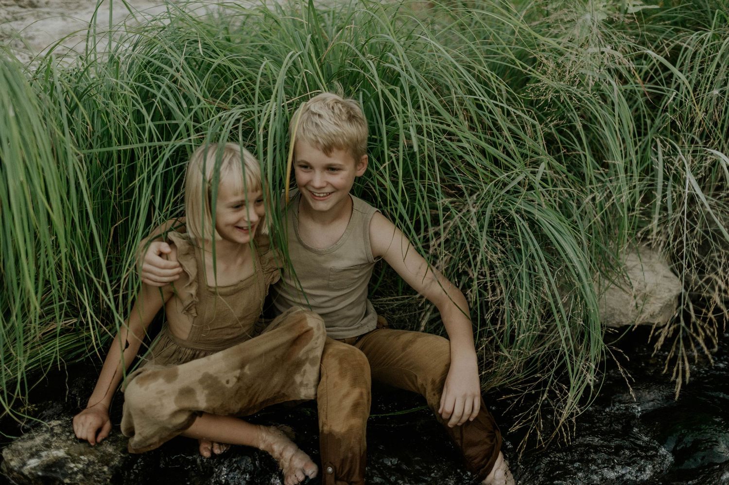 Two muddy blonde children laugh and play together near tall green grass outdoors.