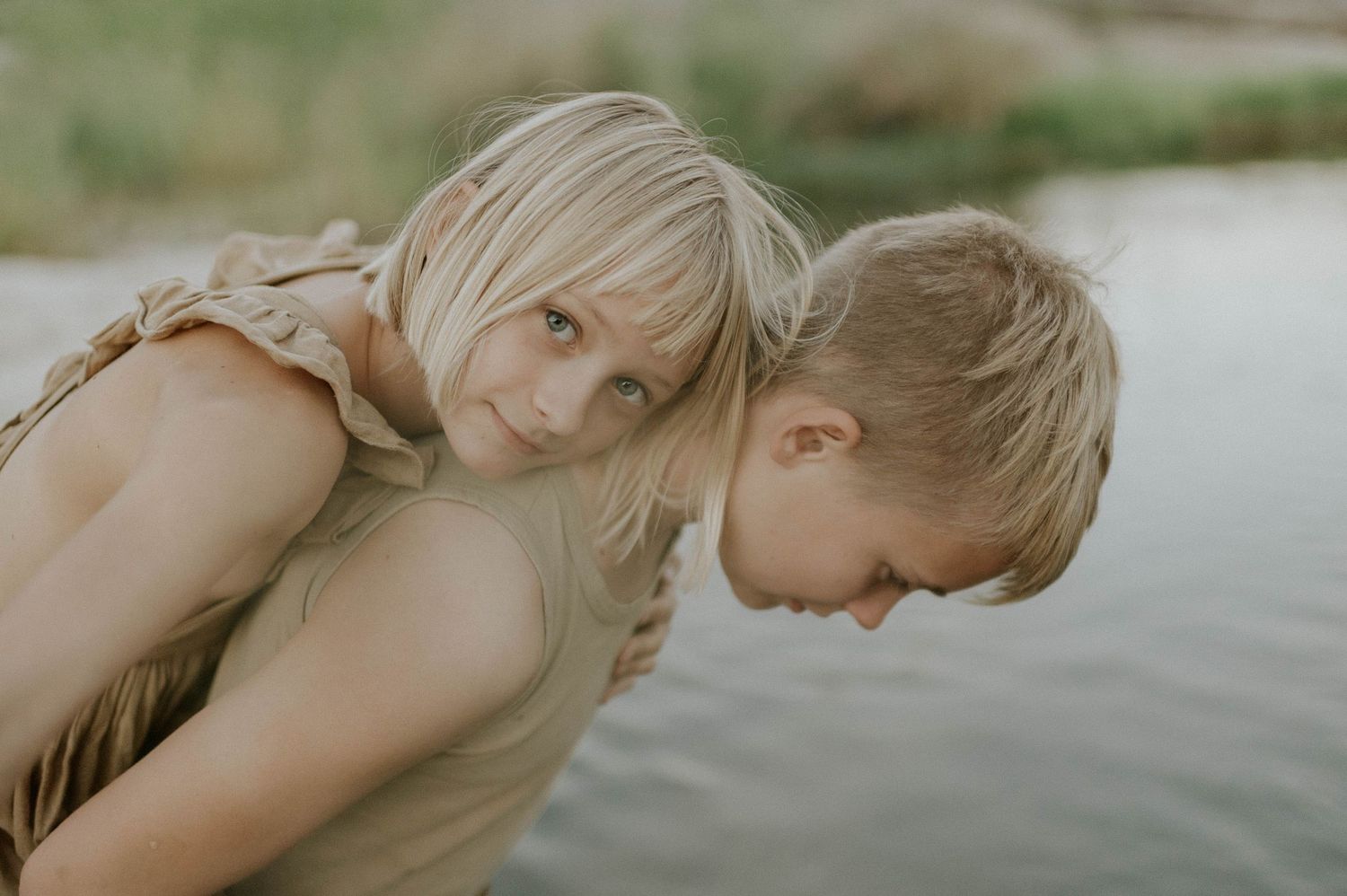 Two blonde children sharing a tender piggyback moment near a serene lake.