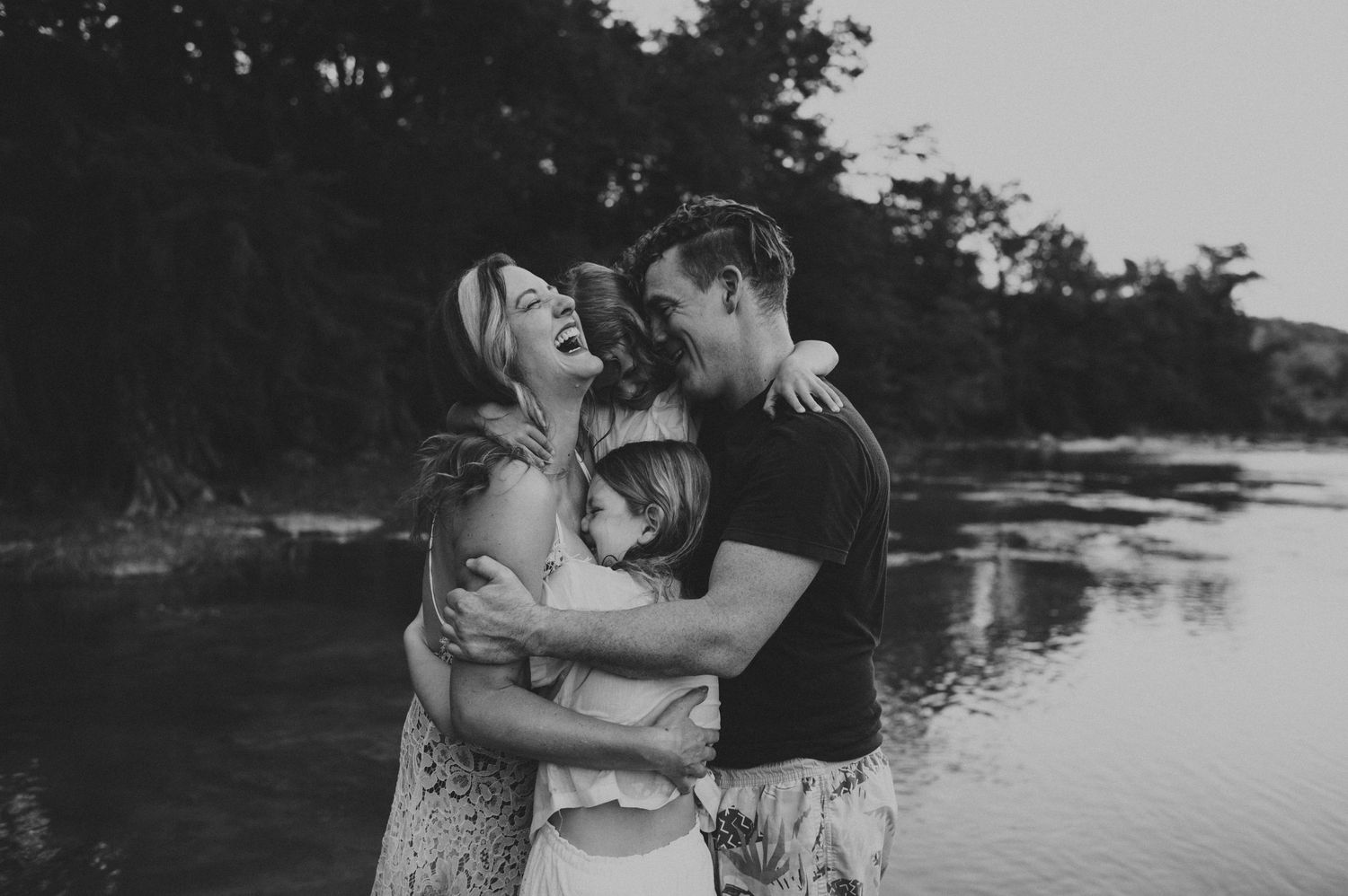 Black and white photo of a happy family embracing and laughing together near a tranquil river surrounded by trees.