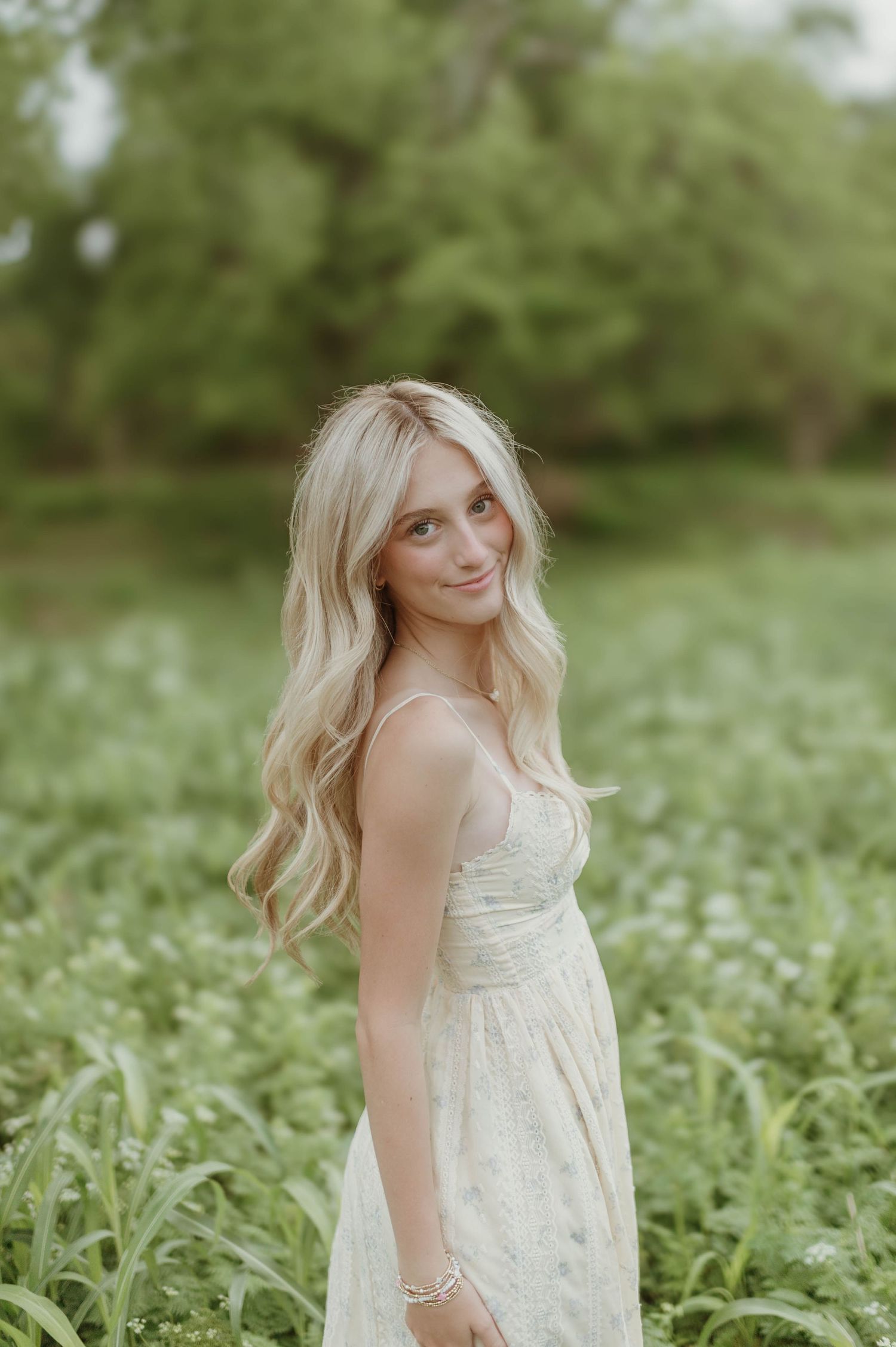 A smiling blonde woman in a white sundress stands in a lush green field of wildflowers.