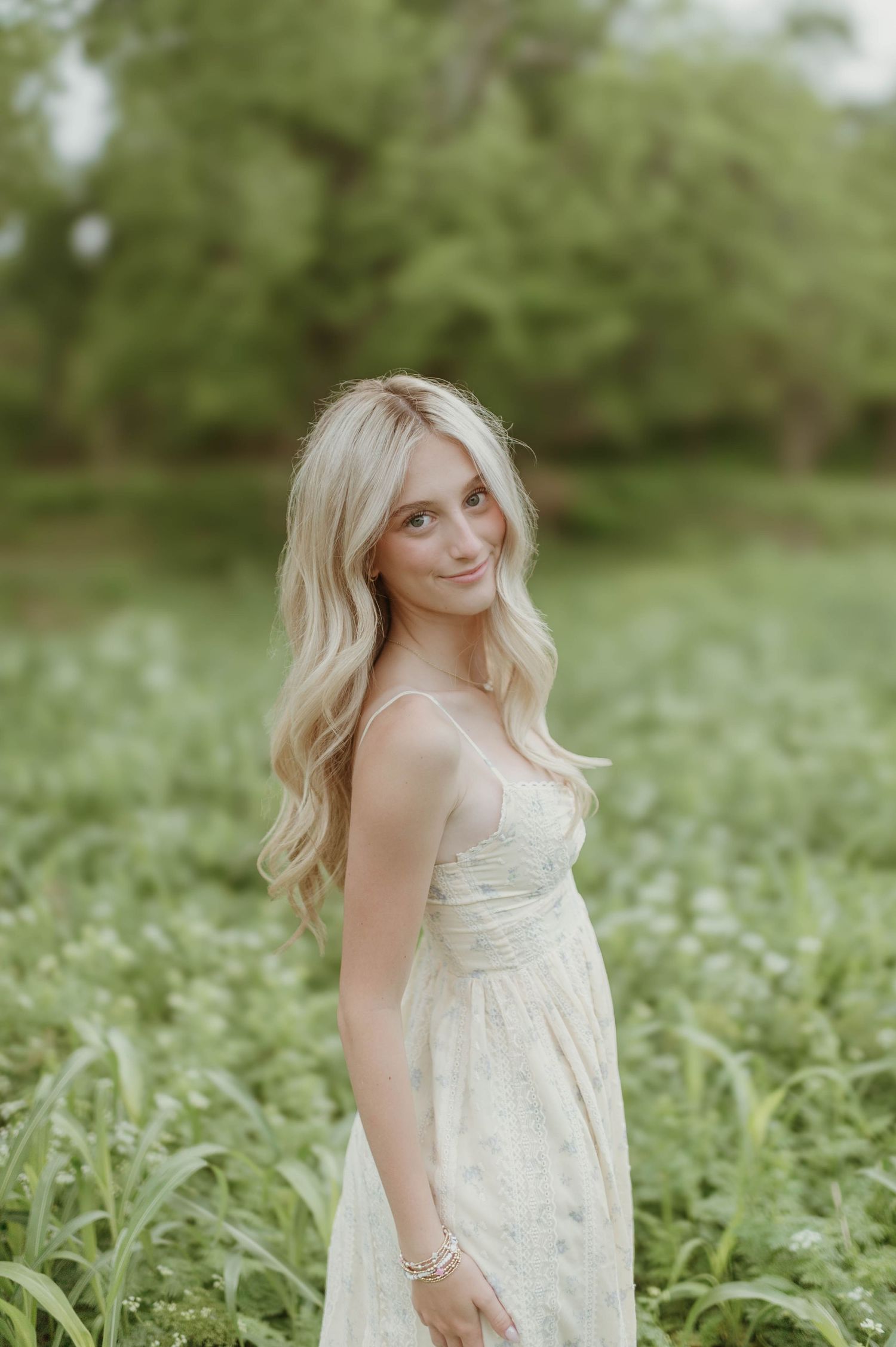 Young blonde woman in white lace dress standing in lush green wildflower field smiling softly.