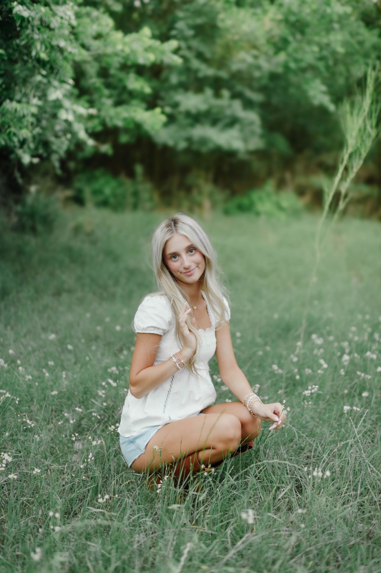 Young blonde woman in white top and denim shorts sitting cross-legged in a lush green meadow with wildflowers.