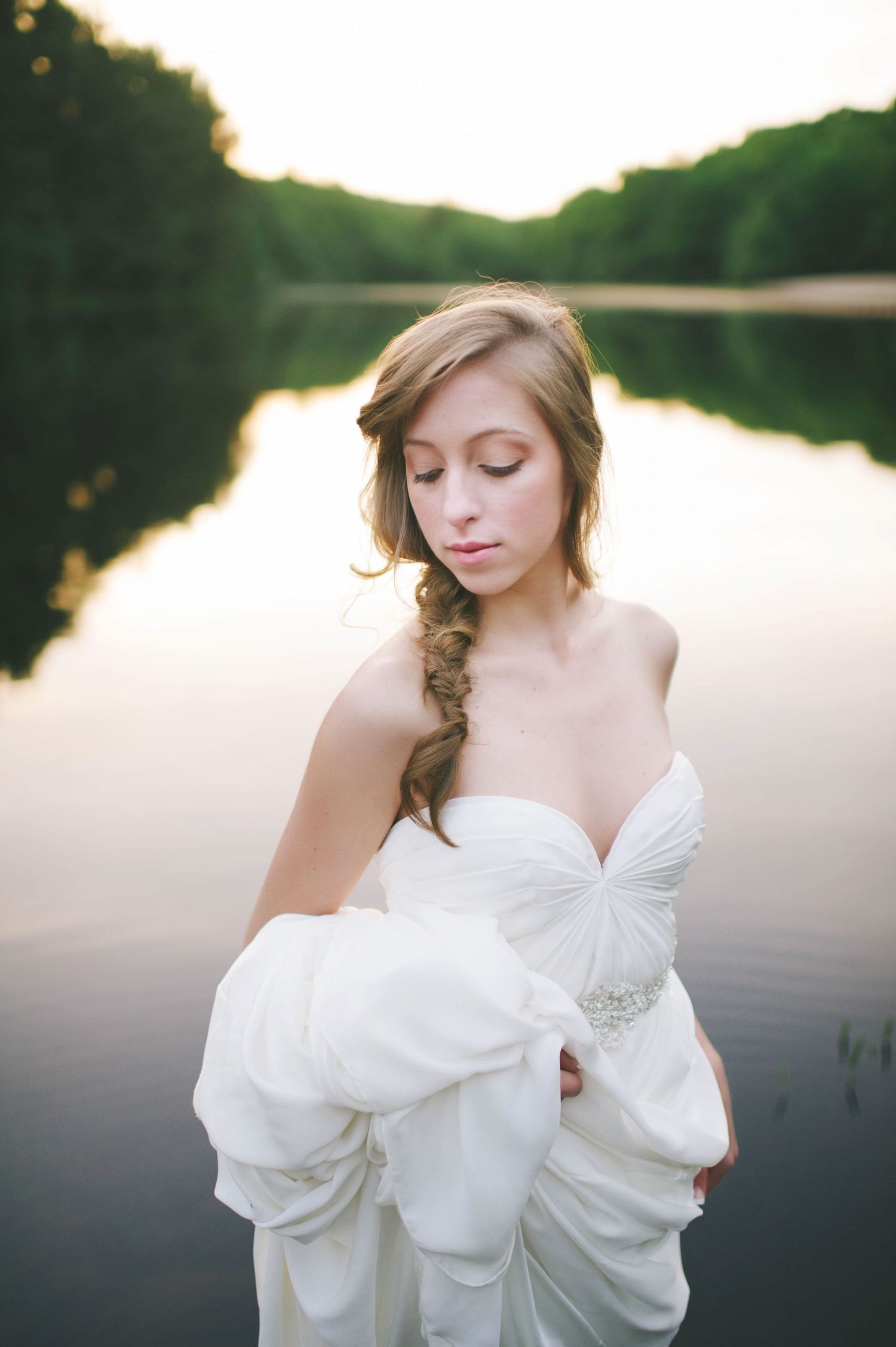 Elegant bride in strapless white gown with braided hair holds wrap beside tranquil sunset lake.