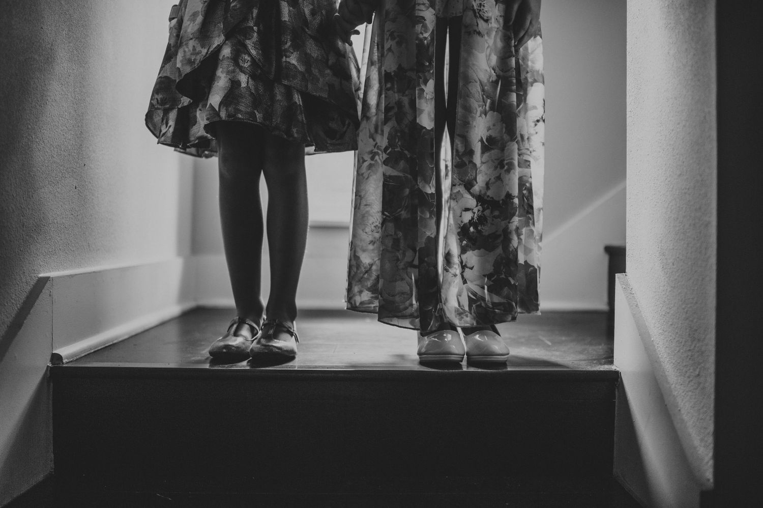Black and white photo of two girls in floral dresses standing on staircase steps.