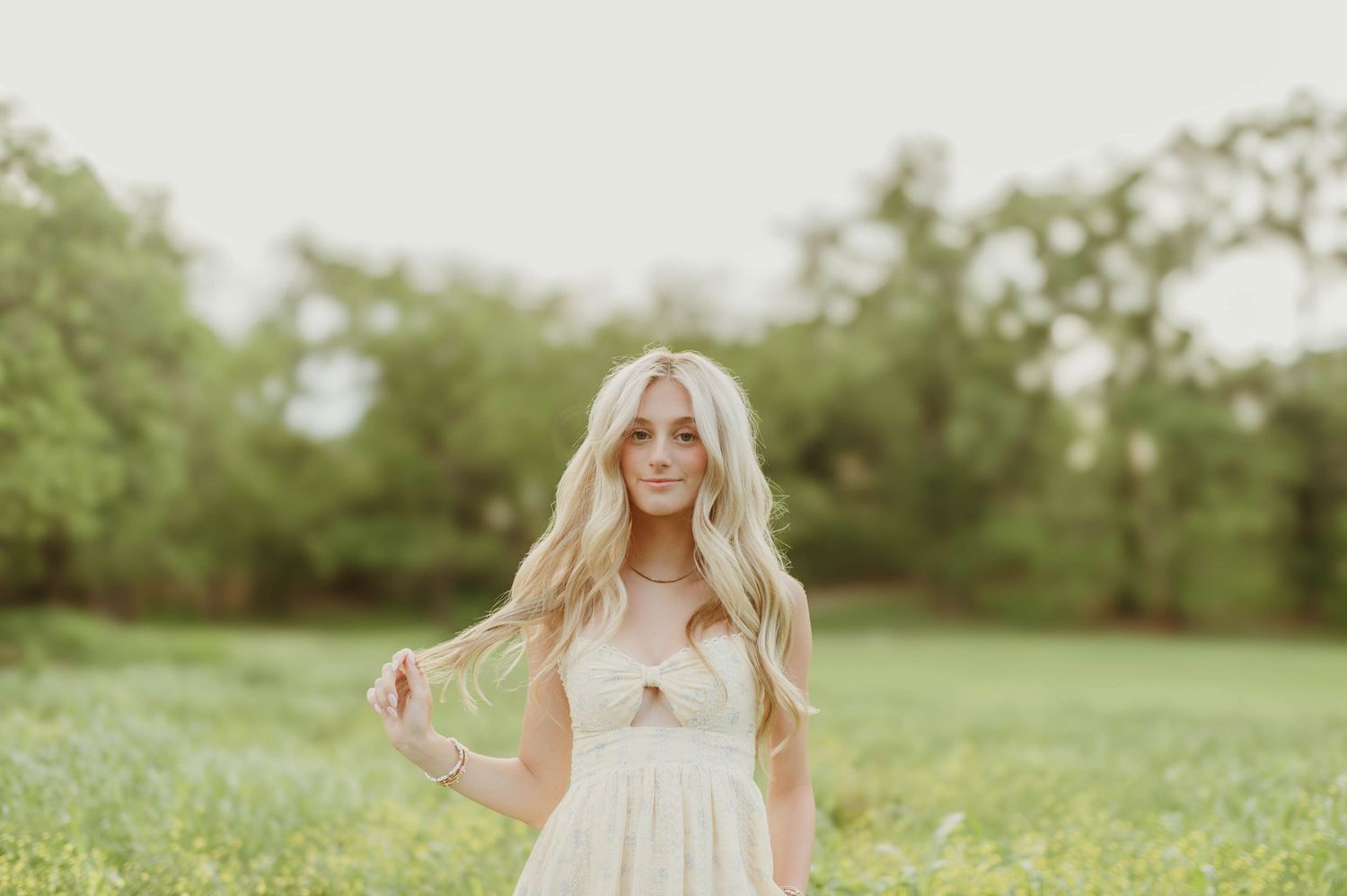 Blonde woman in cream dress standing in green meadow twirling her hair in soft natural light.