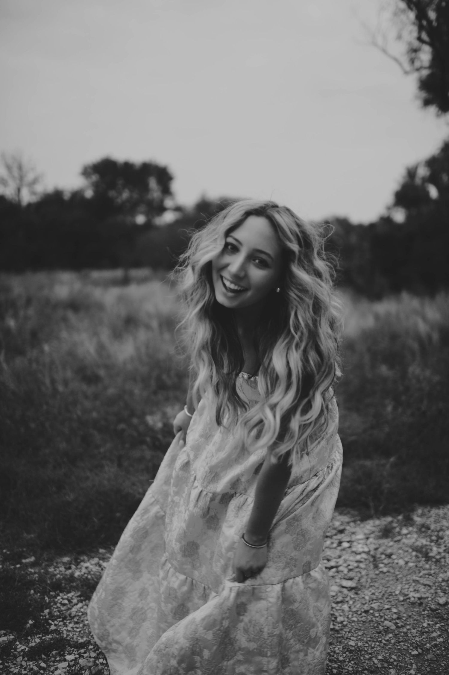 Smiling blonde woman in a flowy white dress poses outdoors in a field, black and white portrait photography.