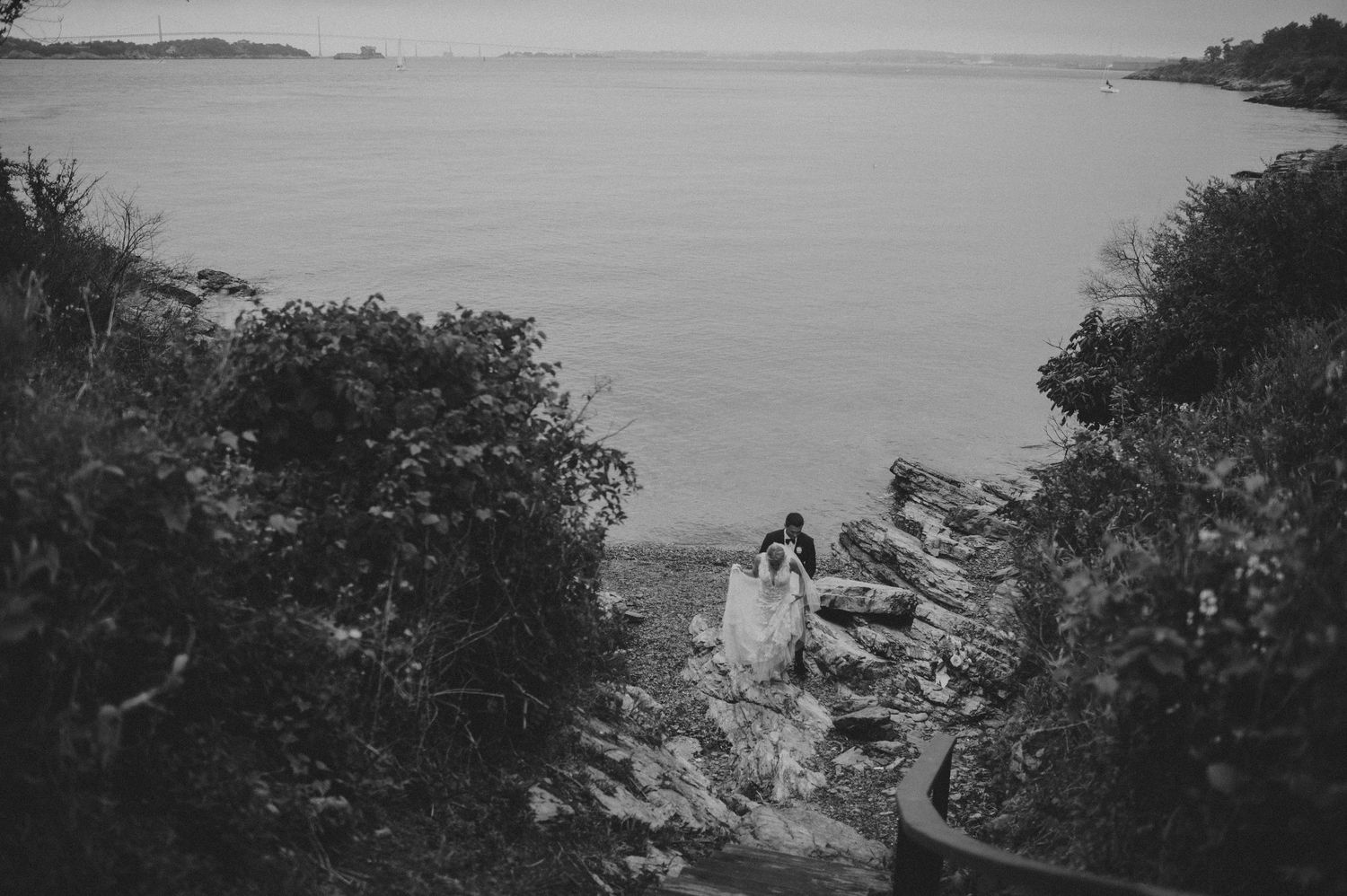 Black and white aerial view of bride and groom sitting on rocky coastal cliffside surrounded by lush greenery.