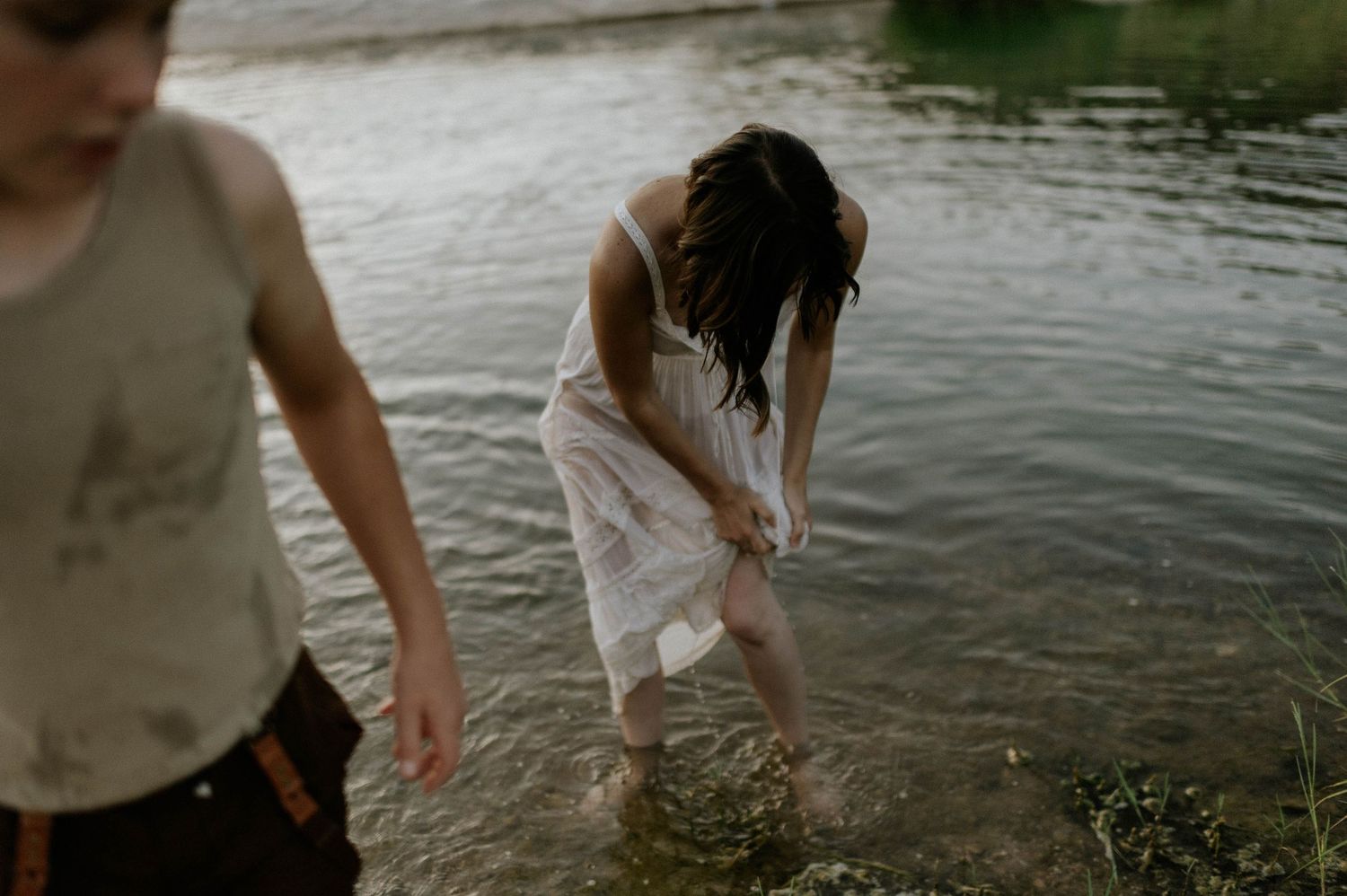 Woman in white dress wading in a river while a child walks past in foreground.