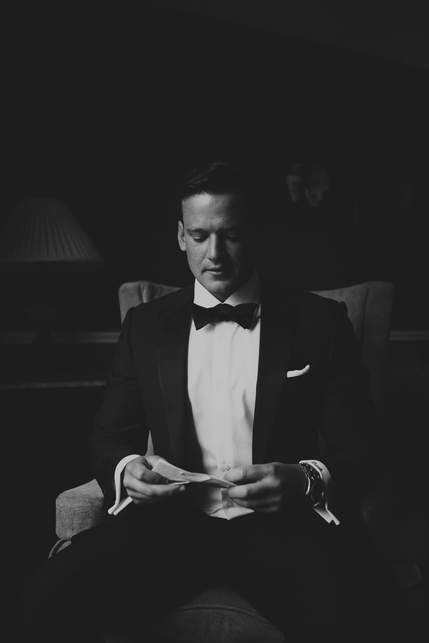 Elegant groom in a black tuxedo and bow tie reading a note while seated in a dimly lit room.