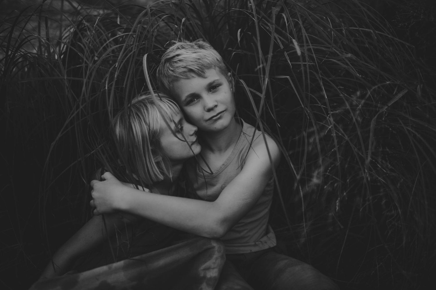 Black and white photo of two children sharing a tender embrace among tall grass outdoors.
