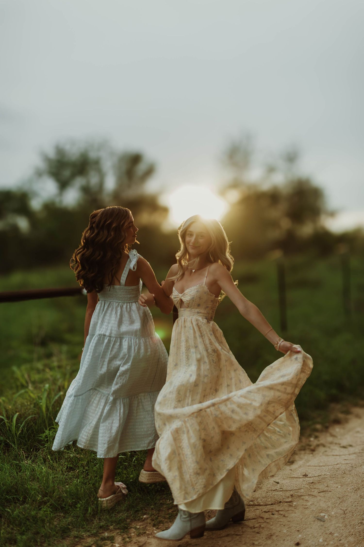 Two women in flowing dresses dancing on a dirt path at golden sunset in a rural setting.