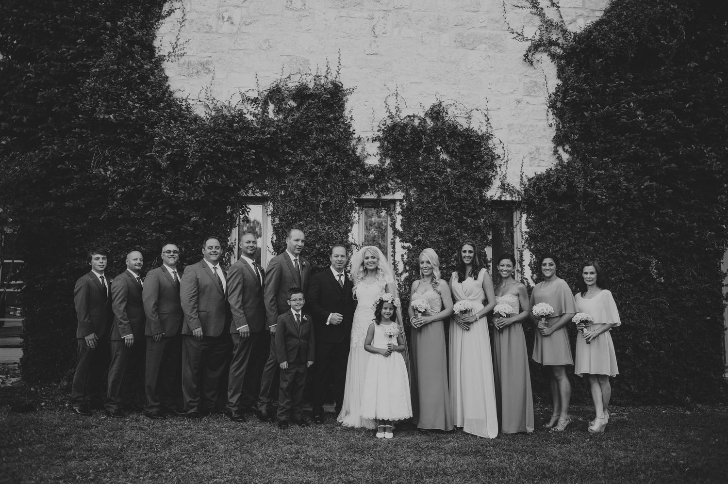 Wedding party poses together outdoors in front of lush ivy-covered backdrop.