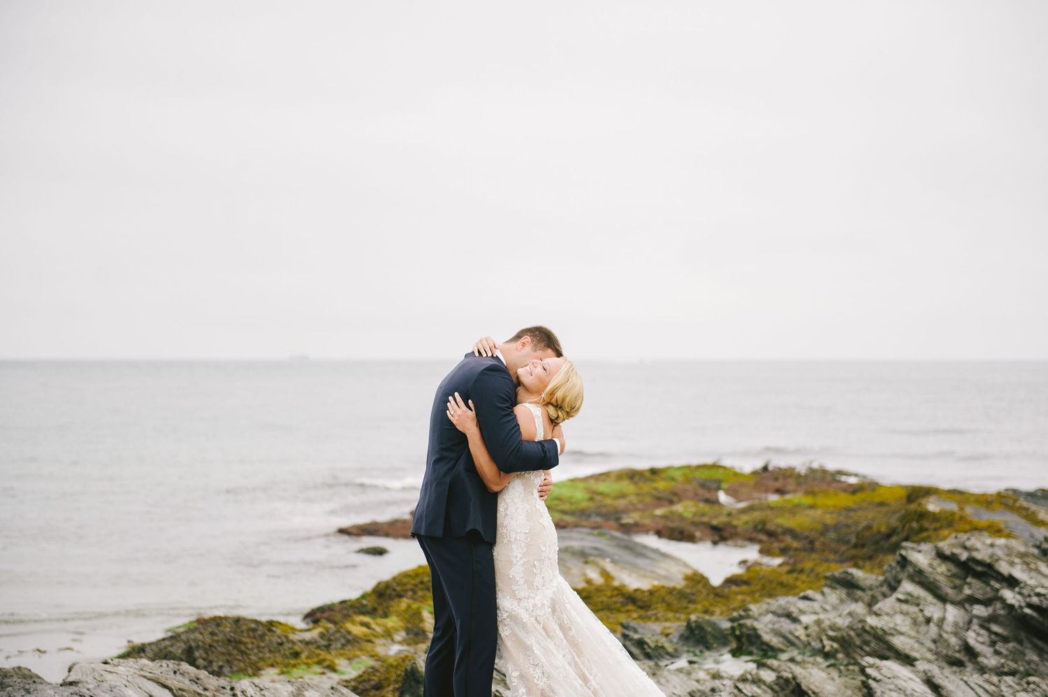 Bride and groom share a romantic embrace on rocky coastal shoreline on wedding day.