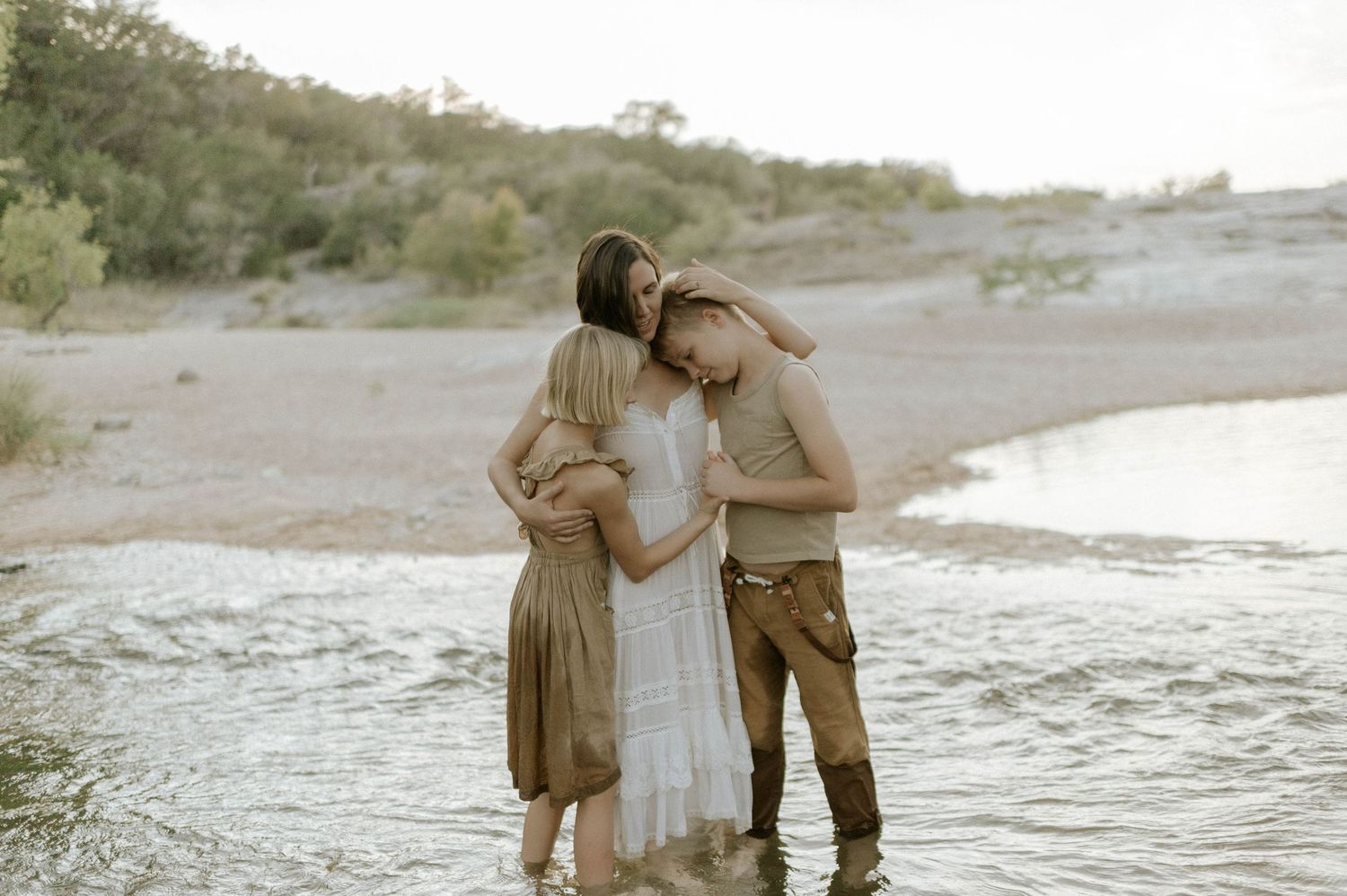 Parents and child share a warm group hug while standing in a shallow river outdoors.