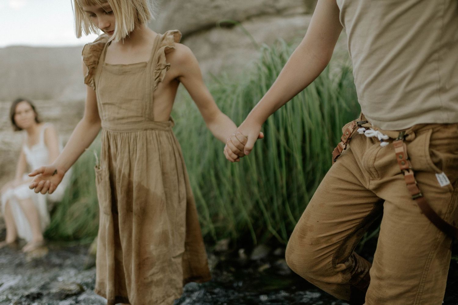 A young girl in a tan dress holds hands with an adult while walking on rocky terrain outdoors.