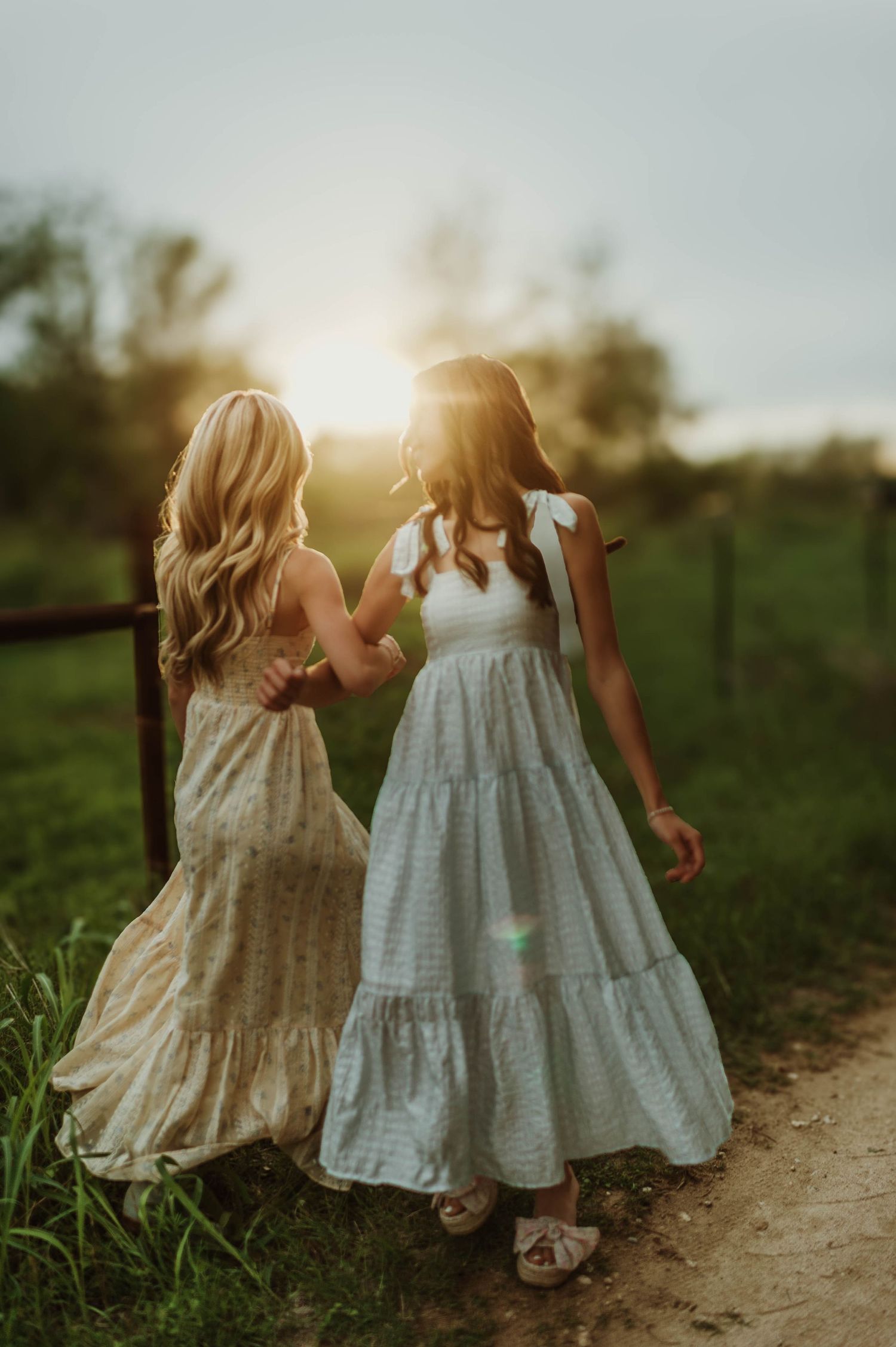 Two young women in flowy maxi dresses hold hands on a dirt path at golden hour sunset.