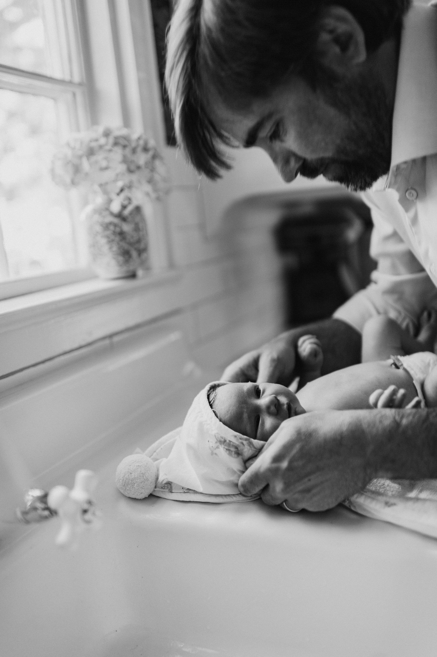 Black and white photo of a father tenderly gazing at his newborn baby lying on a white surface near a window.