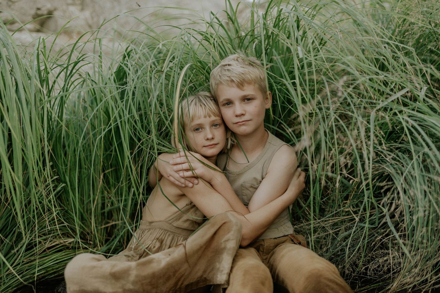 Two blonde children hug warmly while sitting among tall green grass in a nature portrait.